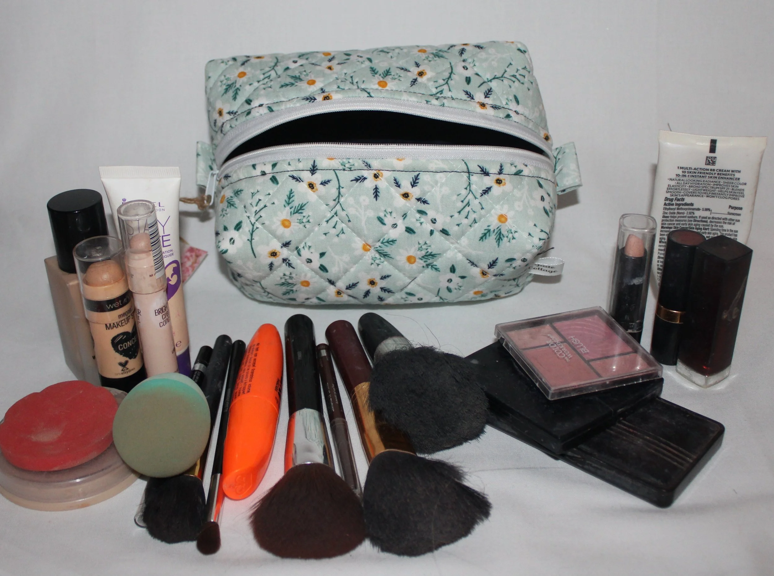 Small Vera travel case in light blue floral quilted print displayed open with the navy lining showing and assorted cosmetics arranged around them.