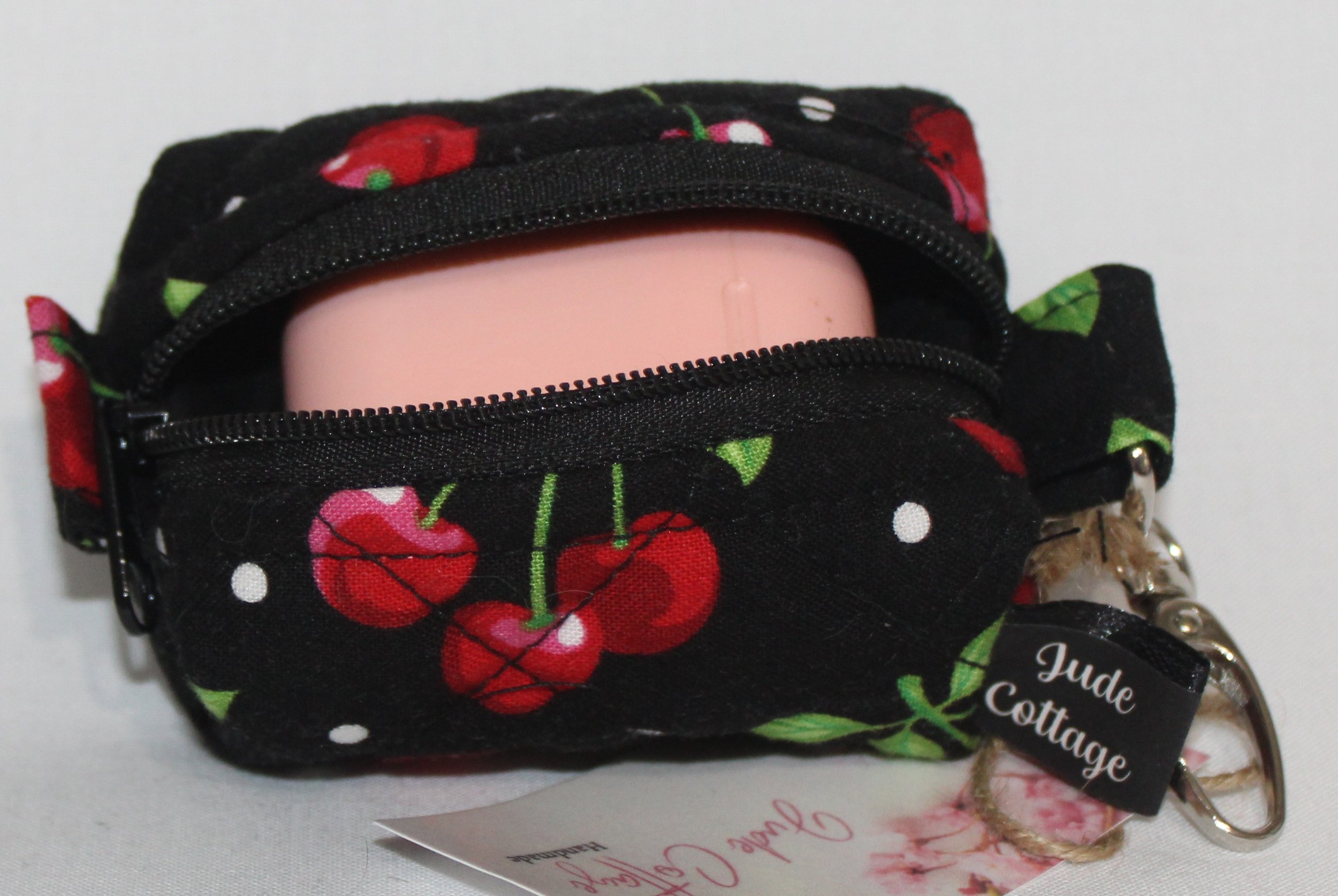 Top open view of the Scattered Cherries Bag Charm showing cherry print on black background with a set of Airpod earbuds inserted for sizing reference.