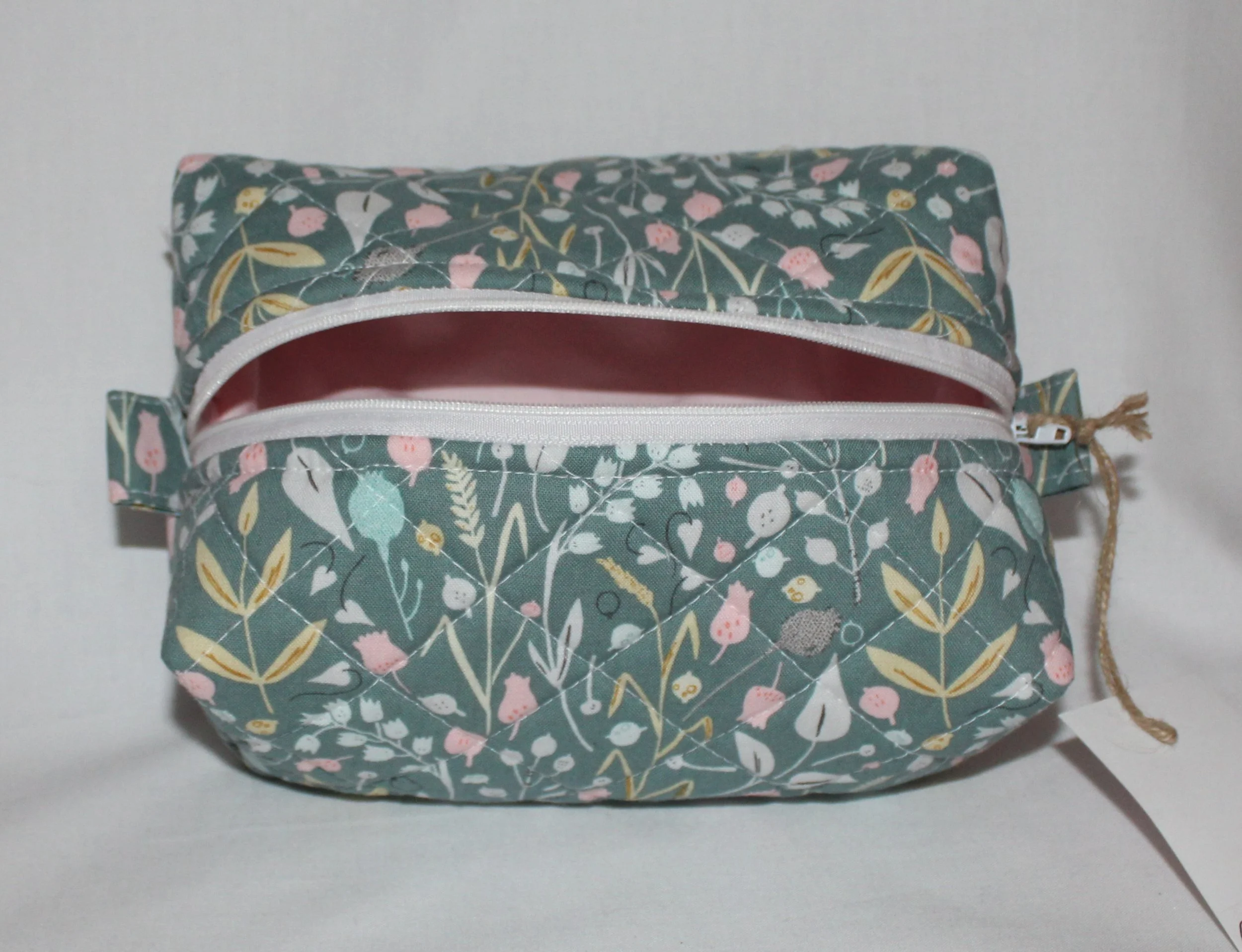 Top‑angle view of the Meadow Breeze quilted travel pouch in green floral fabric with the zipper open showing light pink lining.