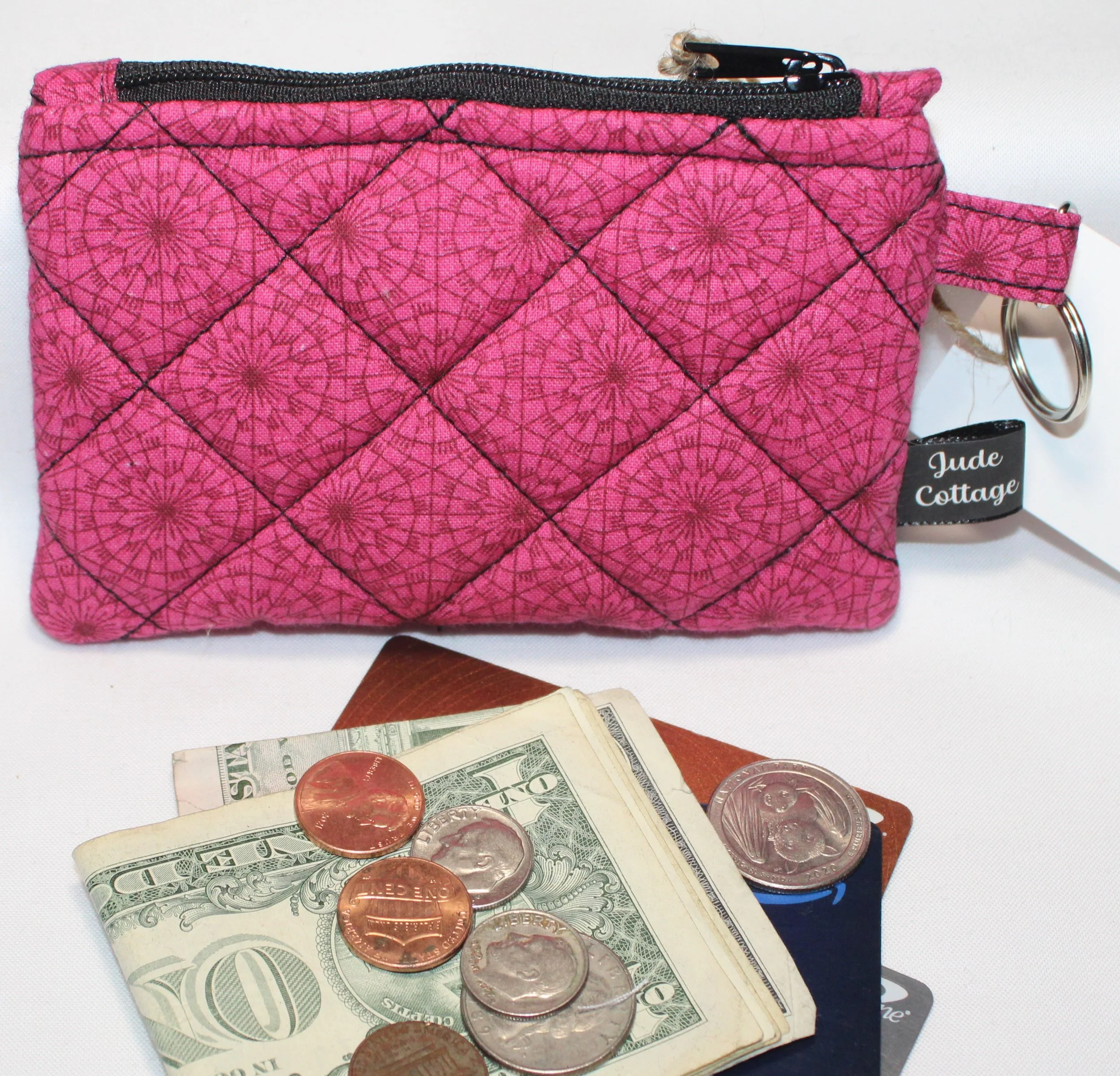 Front view of the Claire quilted fuchsia keychain wallet featuring a geometric background, zipper closure and metal keyring for attachment, shown with cash, coins, and cards to illustrate use.