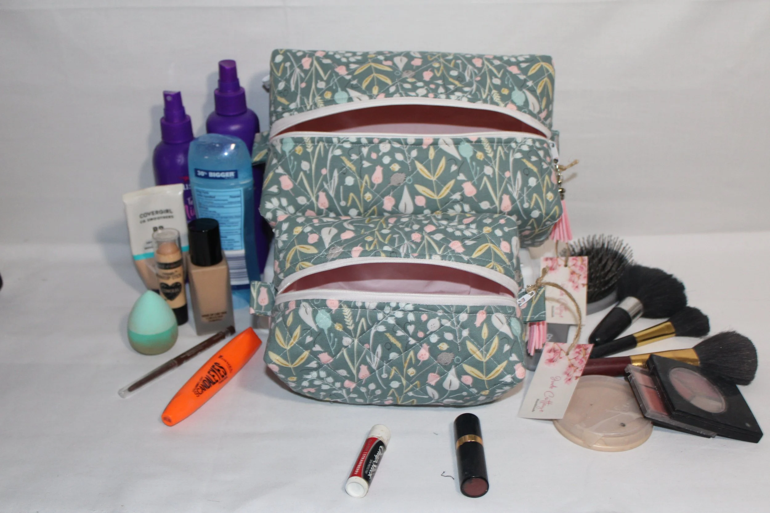 Large and small Meadow Breeze green floral quilted travel pouches shown with makeup, brushes, and personal care items placed around them to illustrate scale.