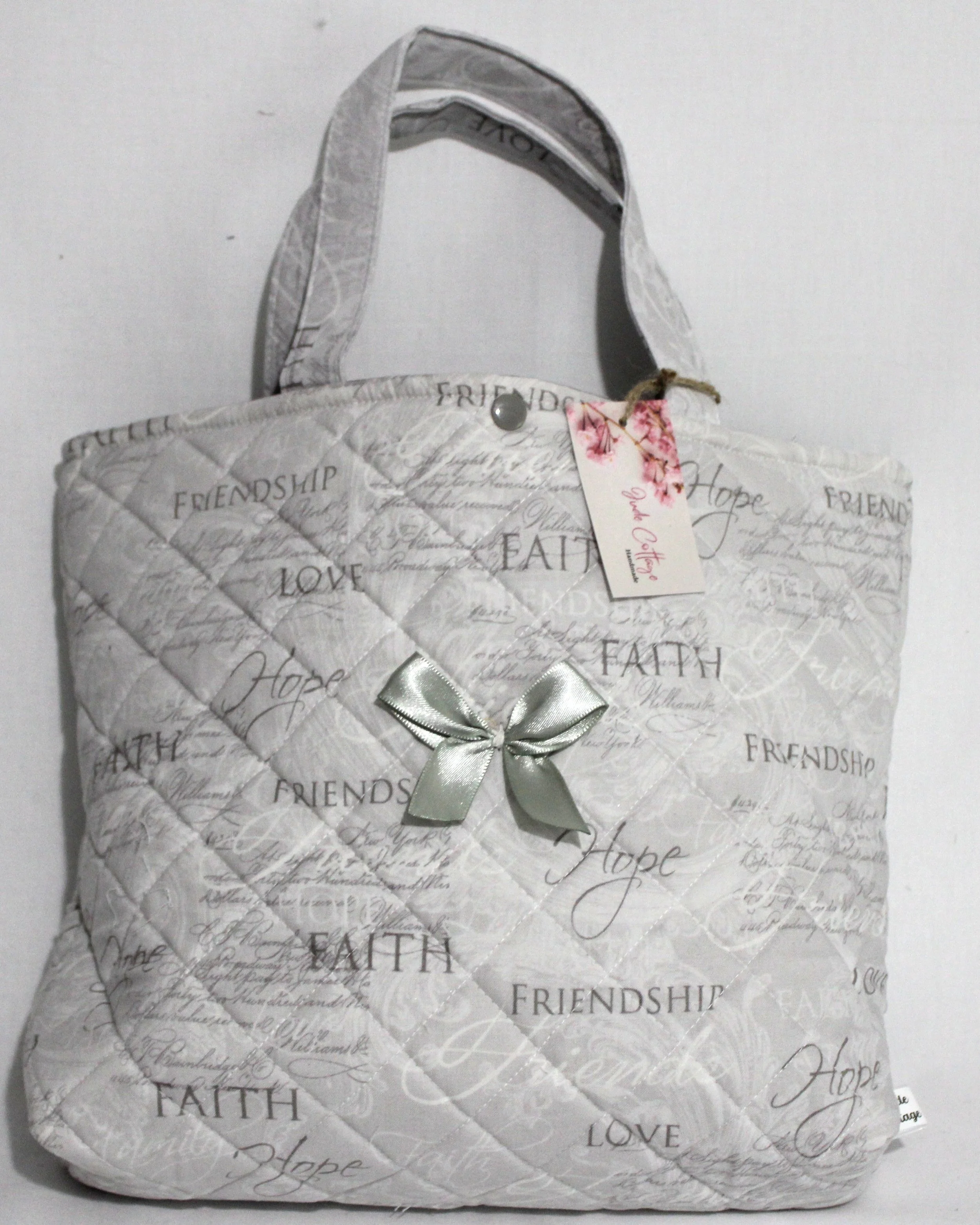 Front view of the quilted Faith Hope Love Bible sleeve with light gray fabric, inspirational wording, and silver bow on front.