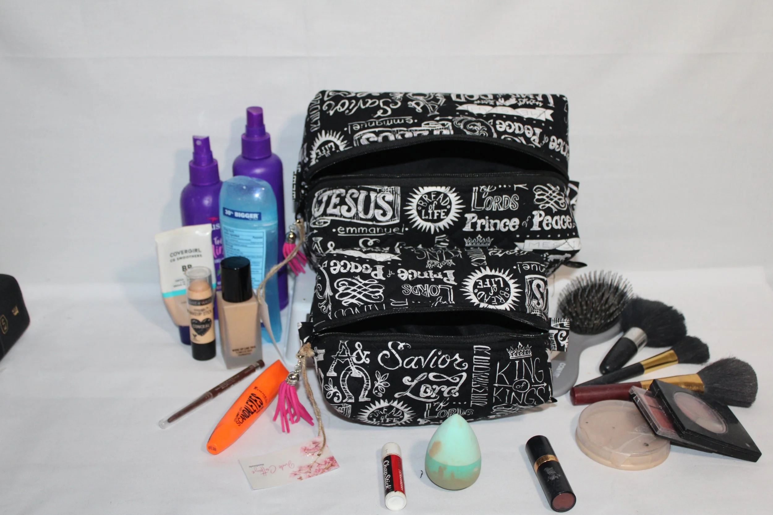 Front view of the large and small black King of Kings travel pouches with white names‑of‑Jesus print, shown with cosmetics and hygiene products arranged in front to illustrate size.