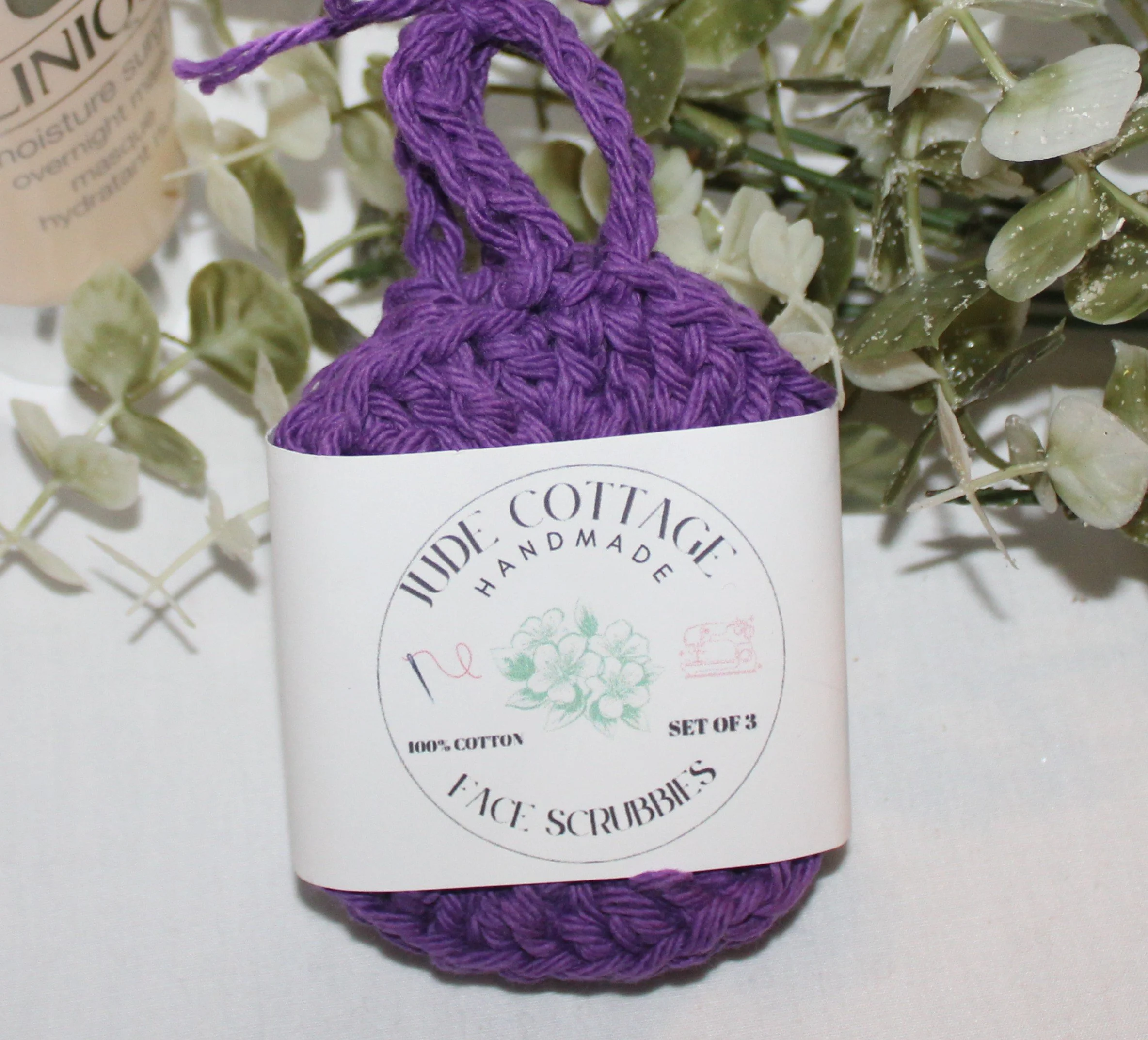 Close‑up of set of three round crocheted plum facial scrubbies with a textured stitch pattern shown with label.
