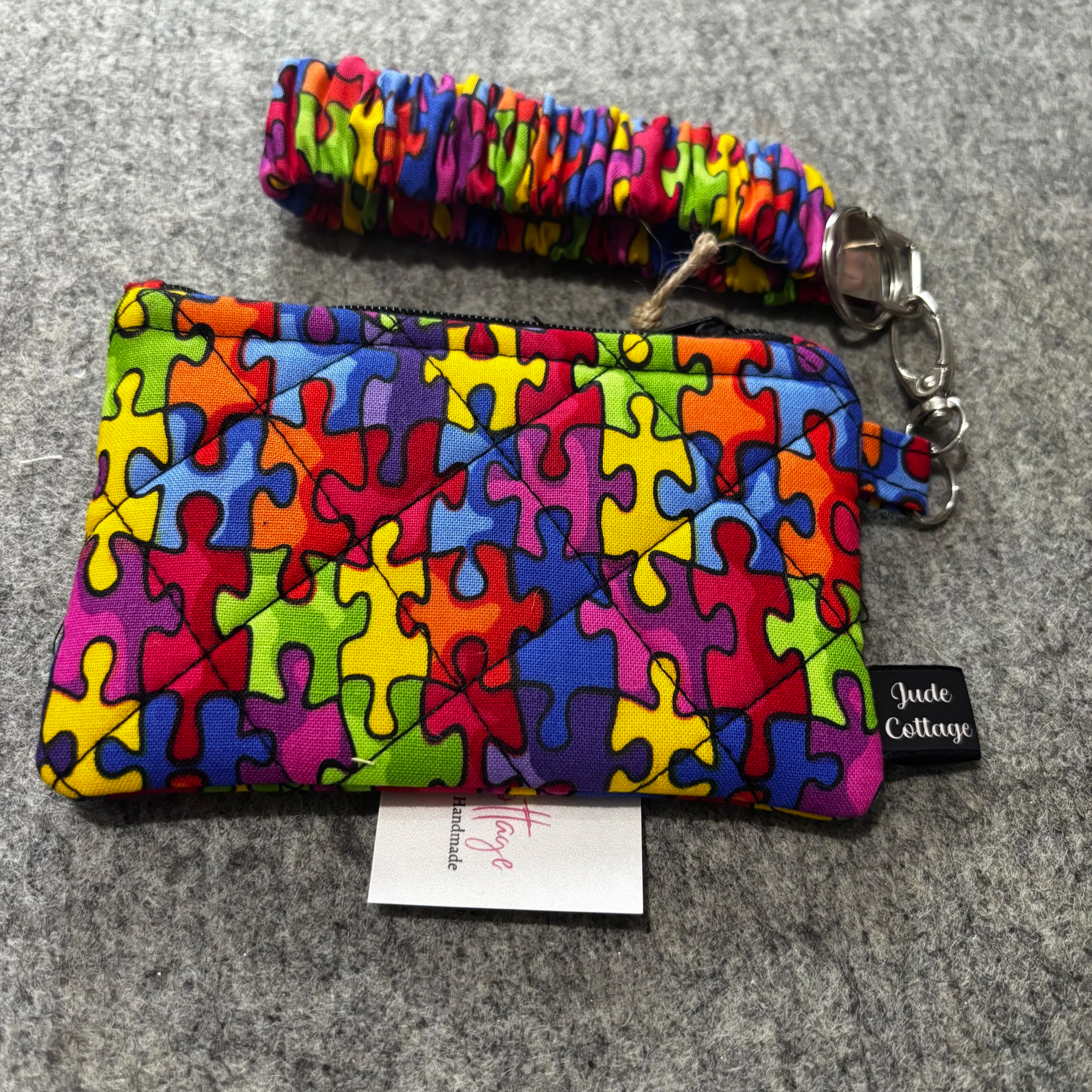 Autism awareness wallet and matching wristlet keychain, both made with a multicolored puzzle‑piece fabric, displayed on a gray surface.