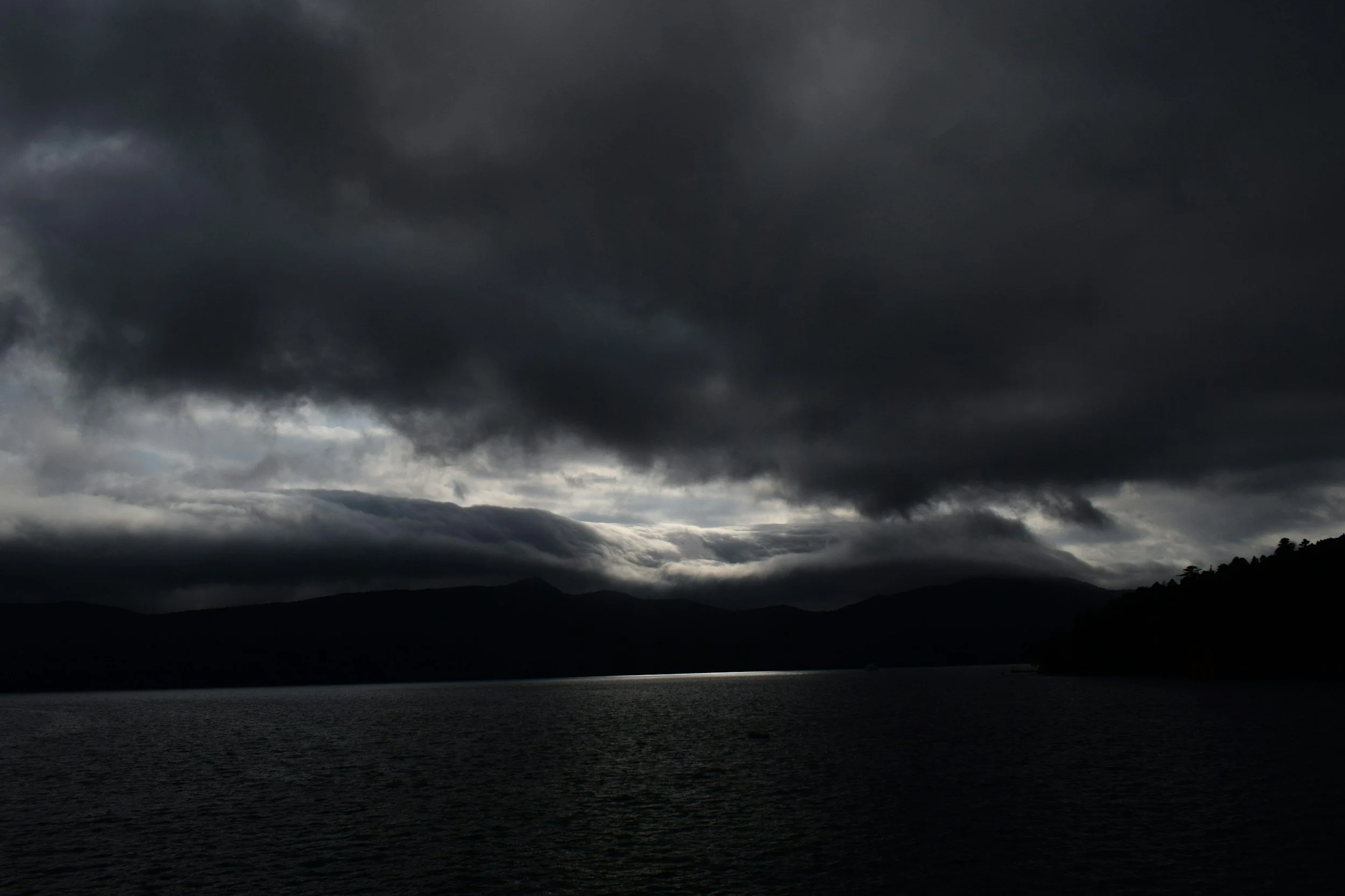 Dark, cloudy sky over a body of water with distant mountains and a silhouetted tree-covered shoreline.