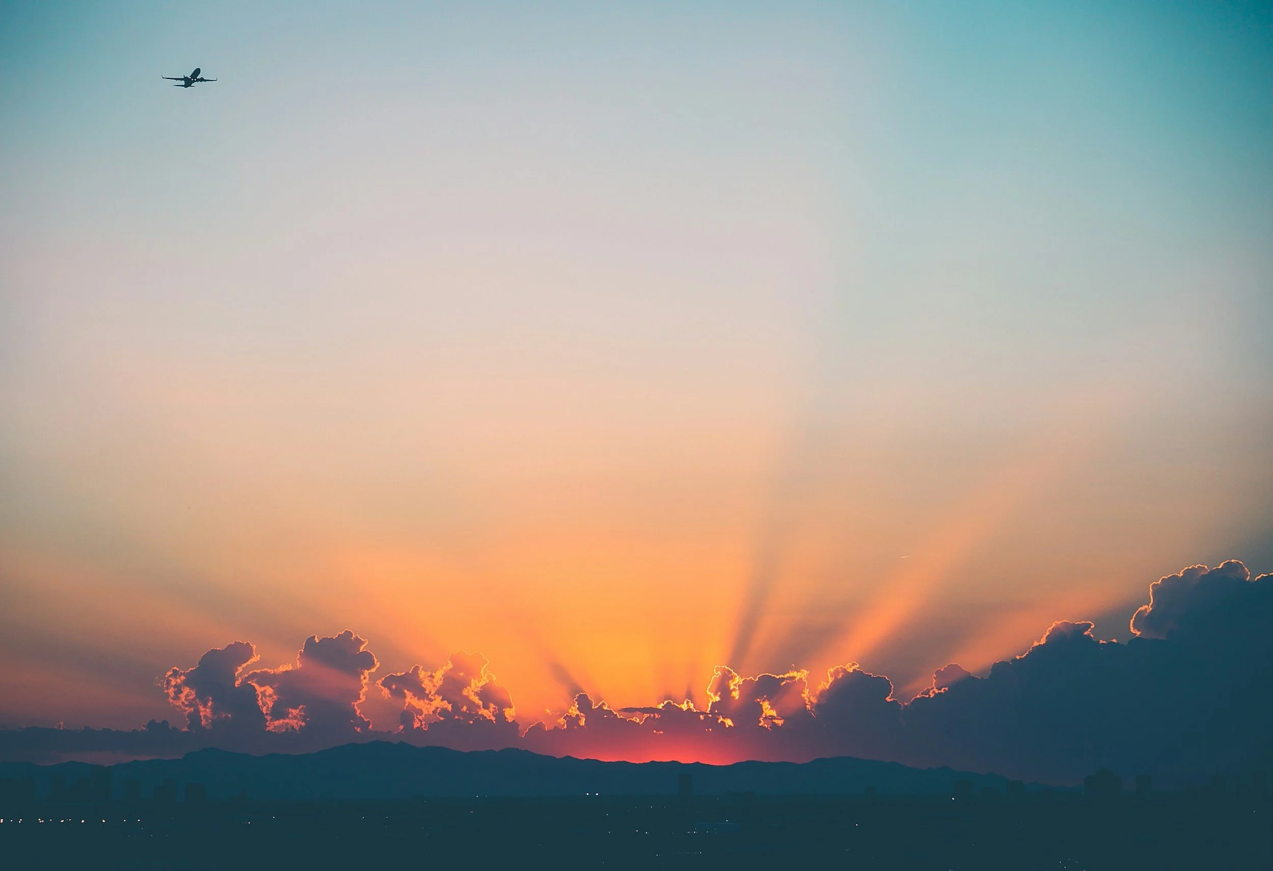 Sunset with orange and pink rays behind dark clouds over mountains. An airplane flying in the sky.
