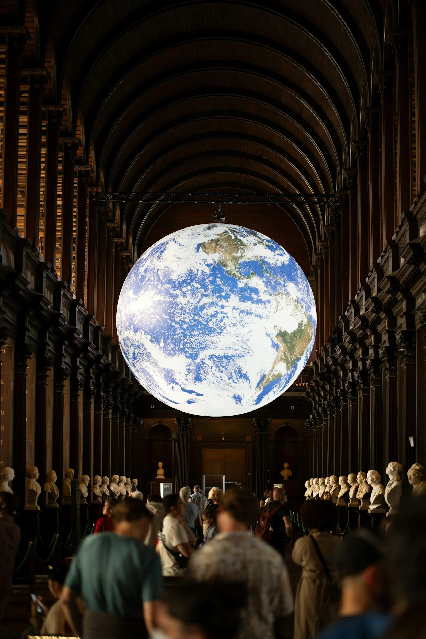 Large globe of Earth hanging from the ceiling of a historic museum hall with high arched wooden ceilings and bust sculptures lining the walls, visitors observing the exhibit.