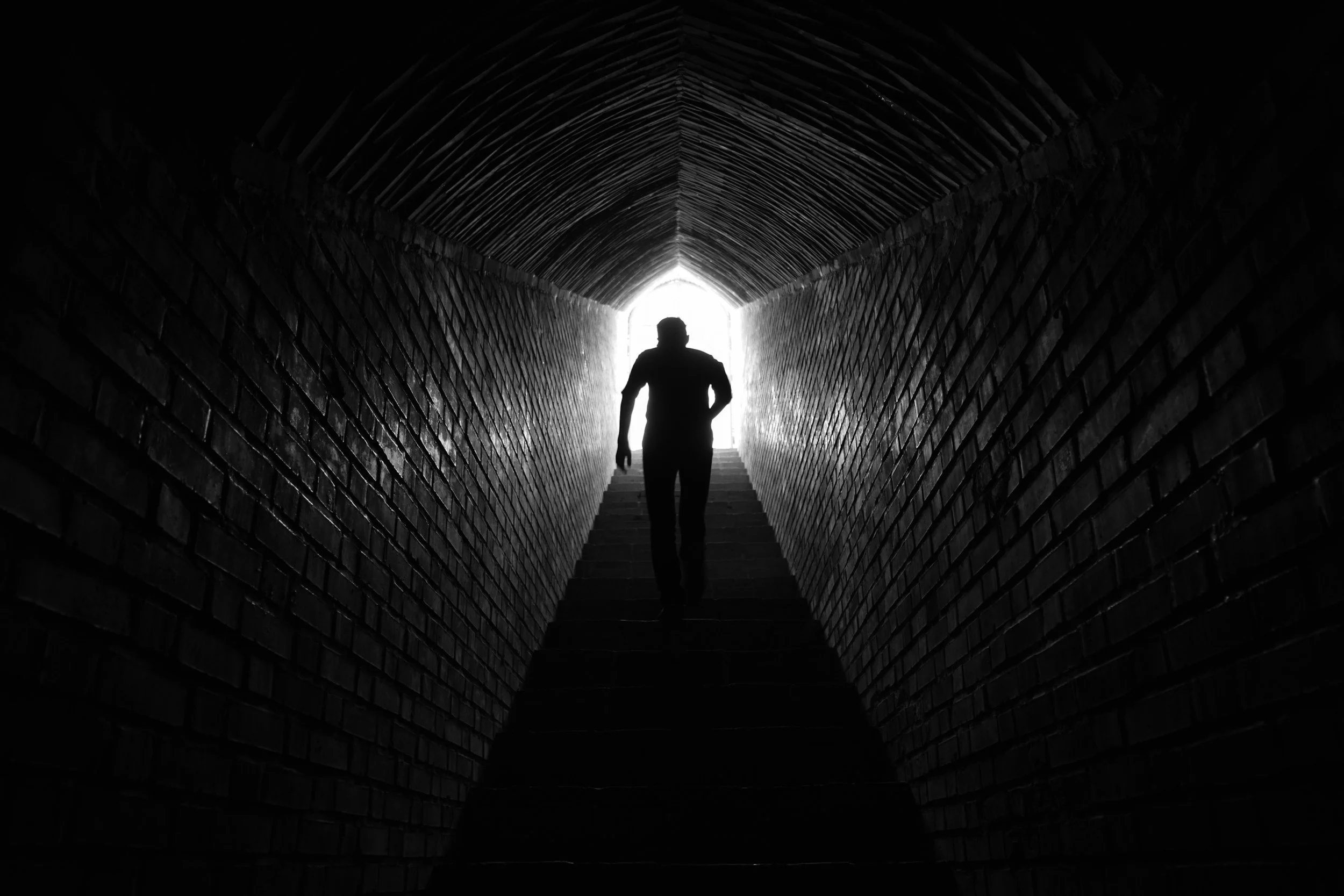 Silhouette of a person walking up a staircase inside a dark brick tunnel towards a bright opening