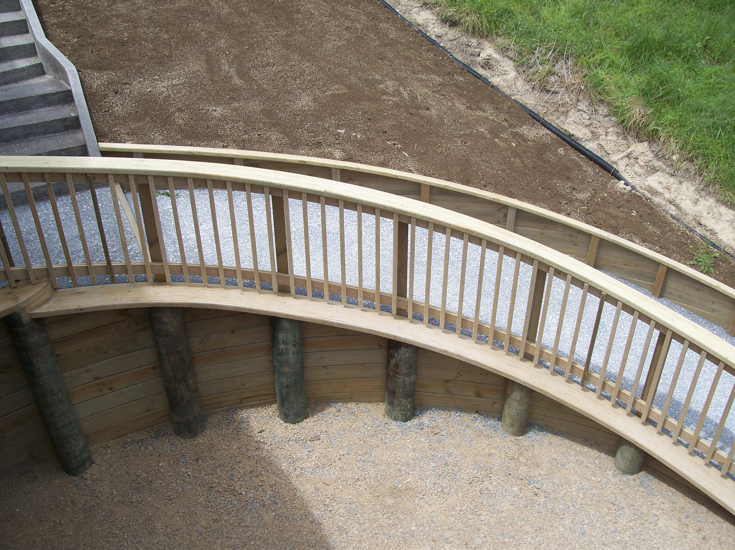 View of a curved wooden staircase with a railing, leading down to a gravel area and a grassy slope in the background.