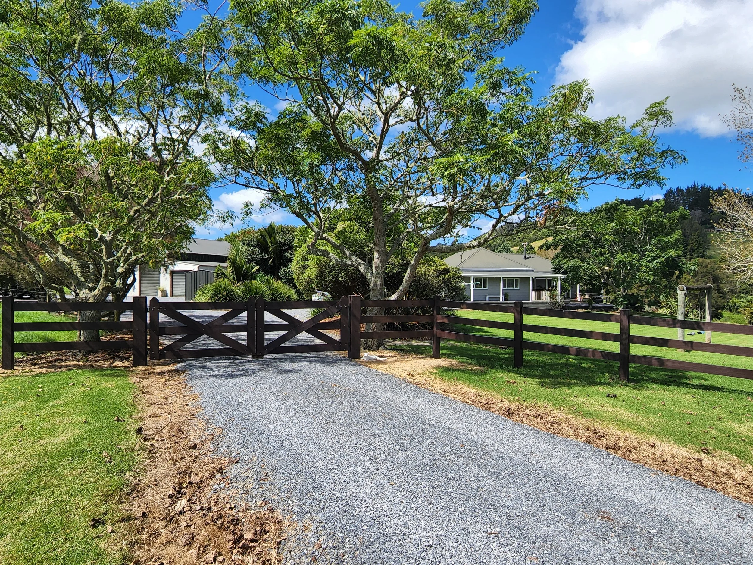 Gravel driveway leading to a closed wooden gate, flanked by fencing and greenery, with trees and a house in the background under a partly cloudy sky.