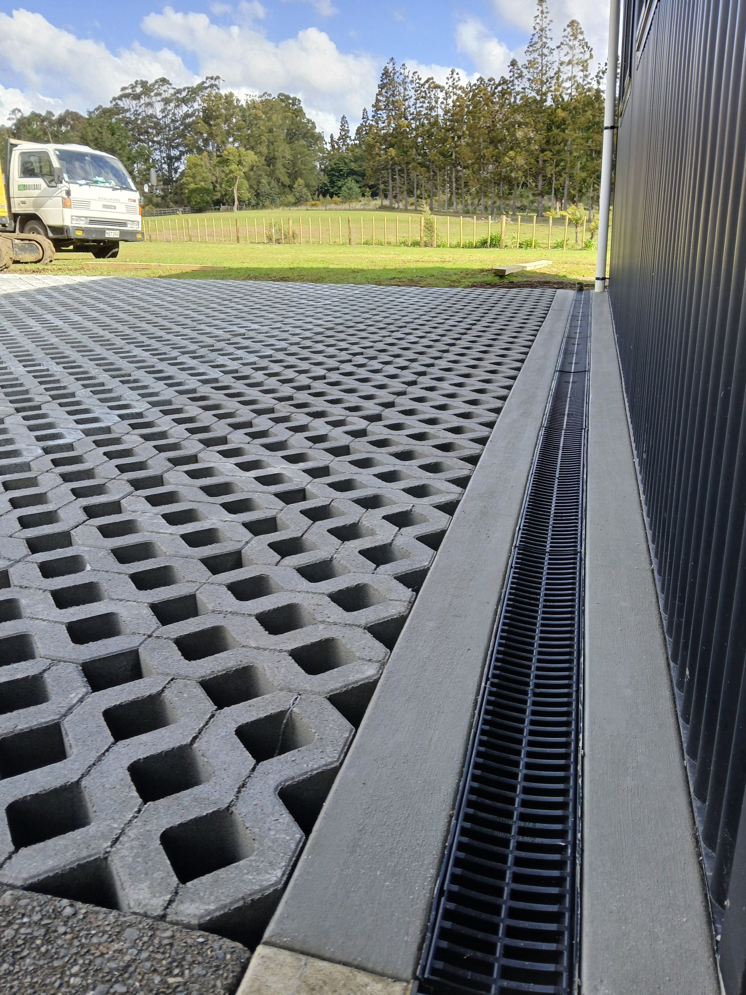 Close-up view of a concrete and plastic drainage system along the base of a building, with grass and trees in the background, and construction vehicle on the left.