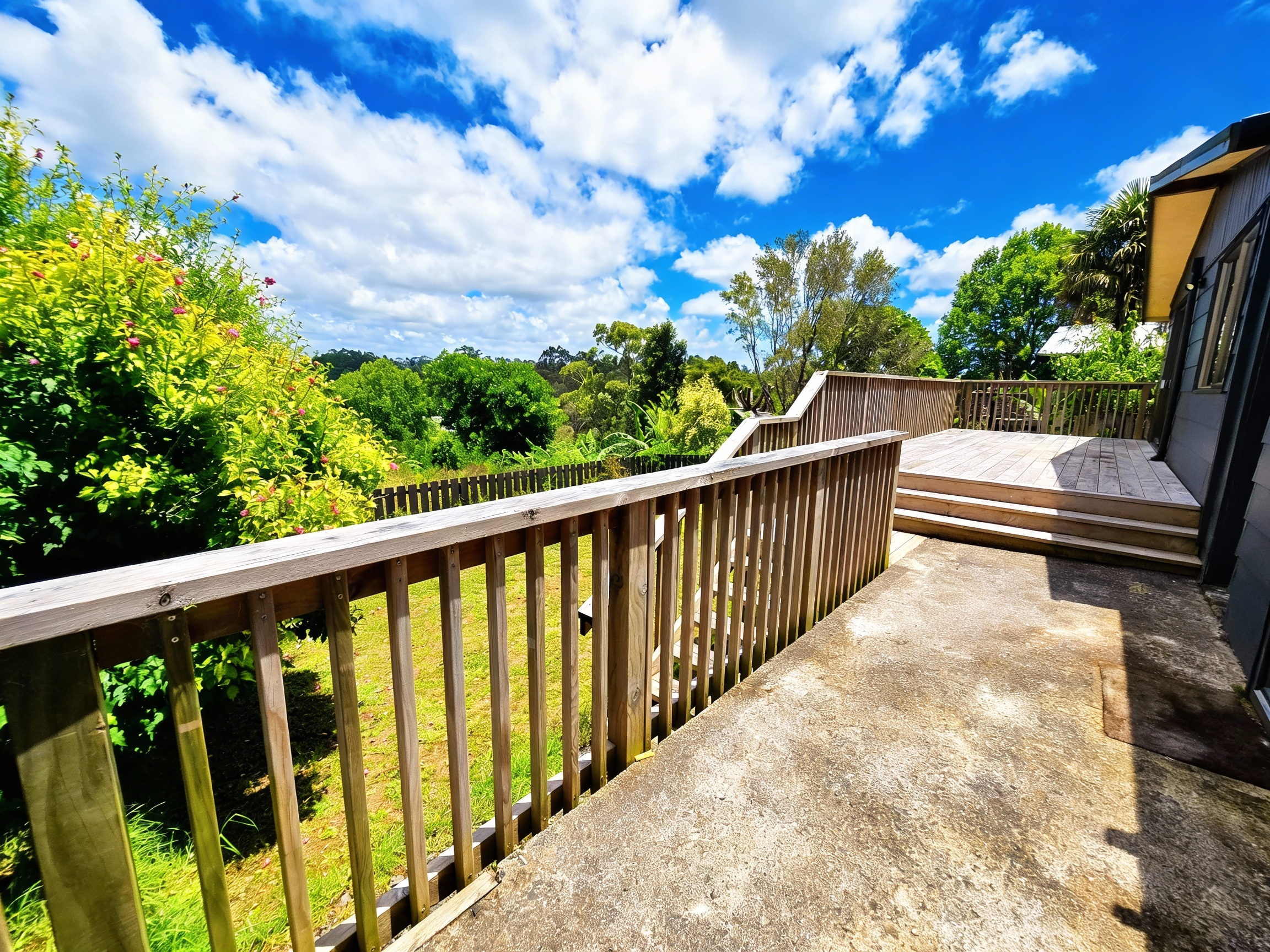 Wooden deck and railing overlooking a green backyard with trees and a partly cloudy blue sky.