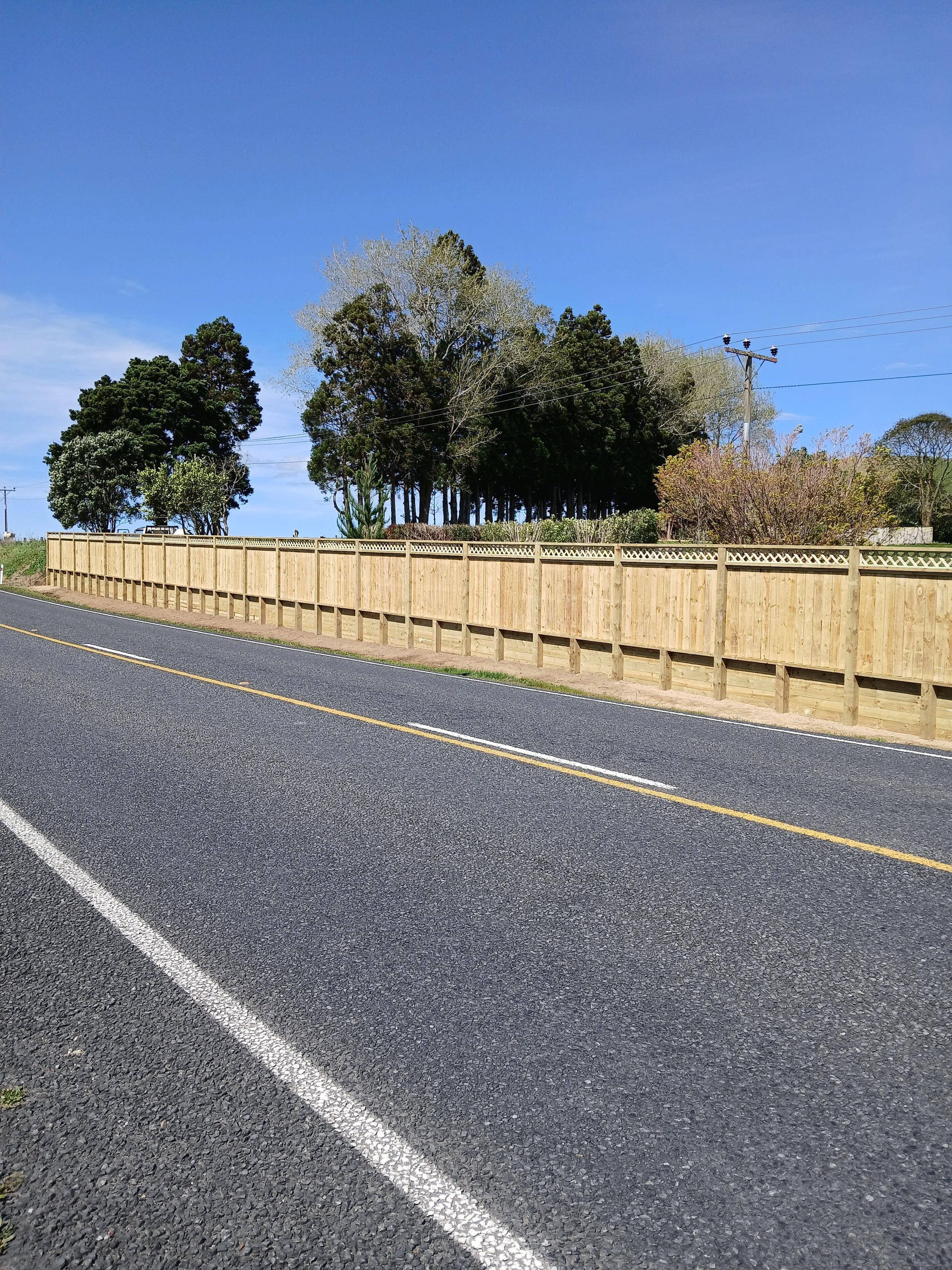 A paved road with double yellow lines, a wooden fence along the side, and trees under a clear blue sky.