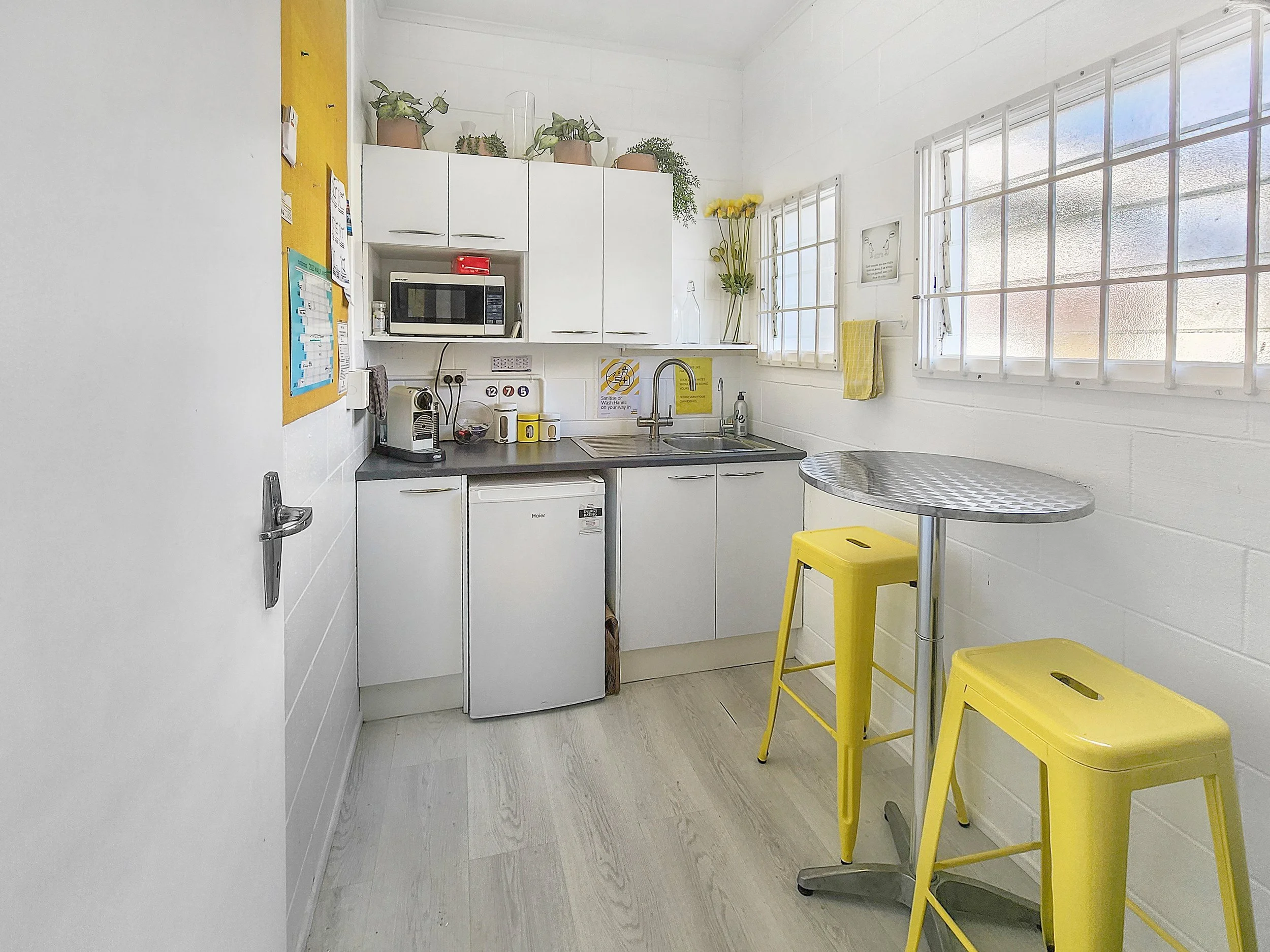 Small kitchen with white cabinets, a microwave, a coffee machine, a mini fridge, a sink, and a yellow stool with a metal table.