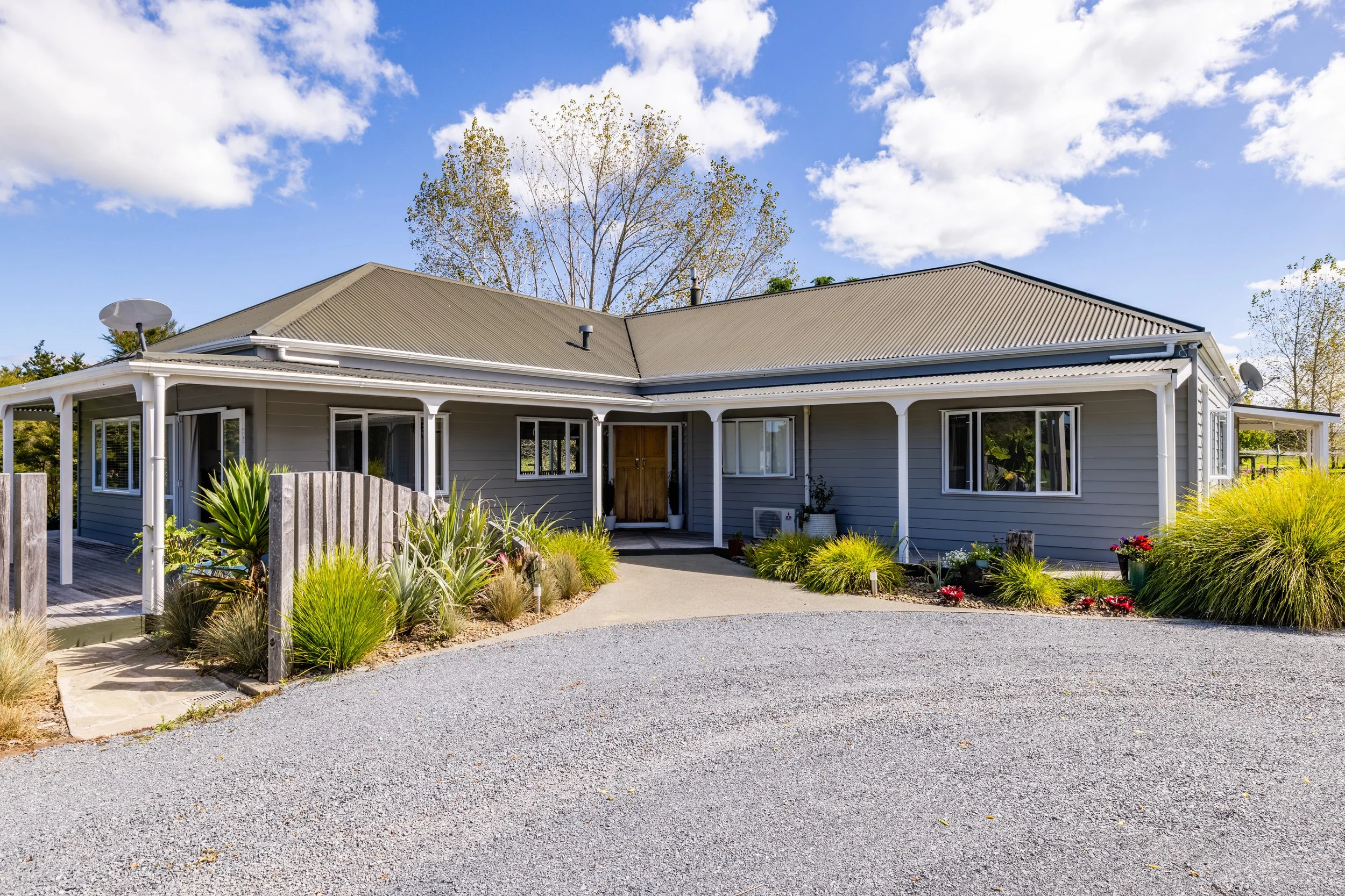 A single-story house with gray siding, white trim, and a metal roof, surrounded by landscaped bushes and plants, with a gravel driveway and a clear blue sky with some clouds.