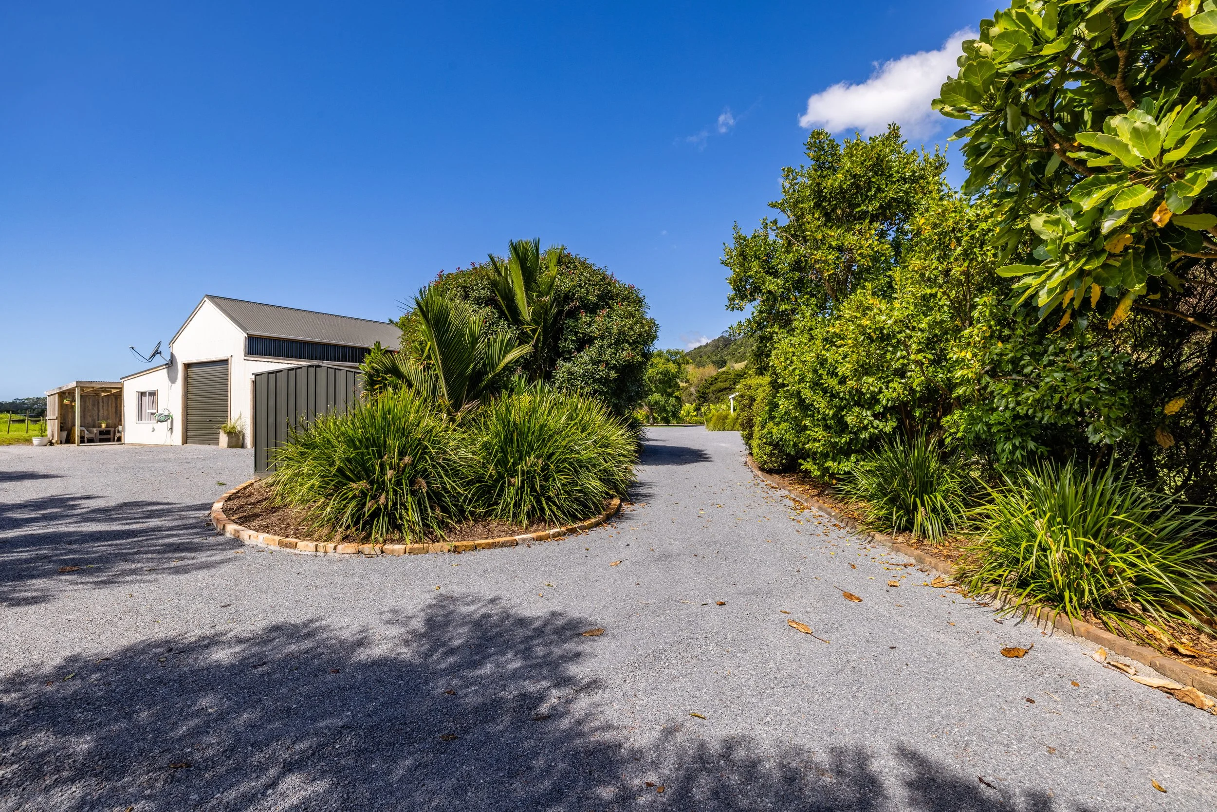 A gravel driveway curves around lush green bushes and trees leading to a white building with a gray roof and roller door, under a bright blue sky.