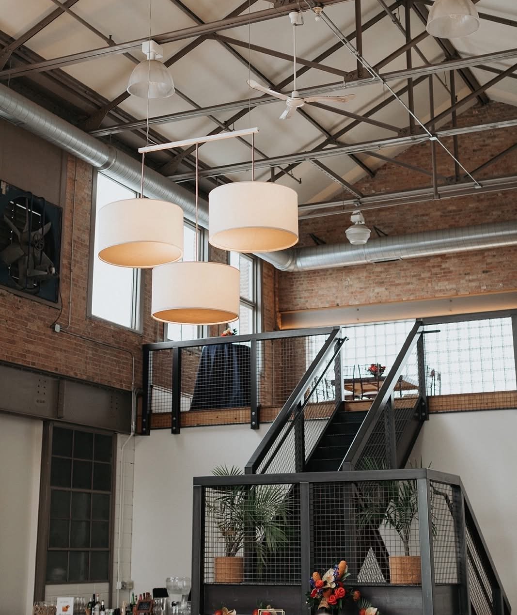 Interior view of a modern industrial-style space with hanging white drum pendant lights, exposed brick walls, metal ductwork, and a staircase leading to a second level.