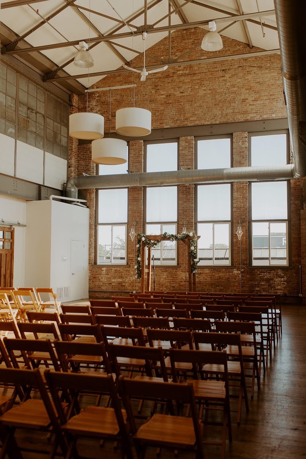 Indoor wedding ceremony setup with rows of wooden chairs facing an arch decorated with greenery and white flowers, large windows, brick walls, and hanging light fixtures.