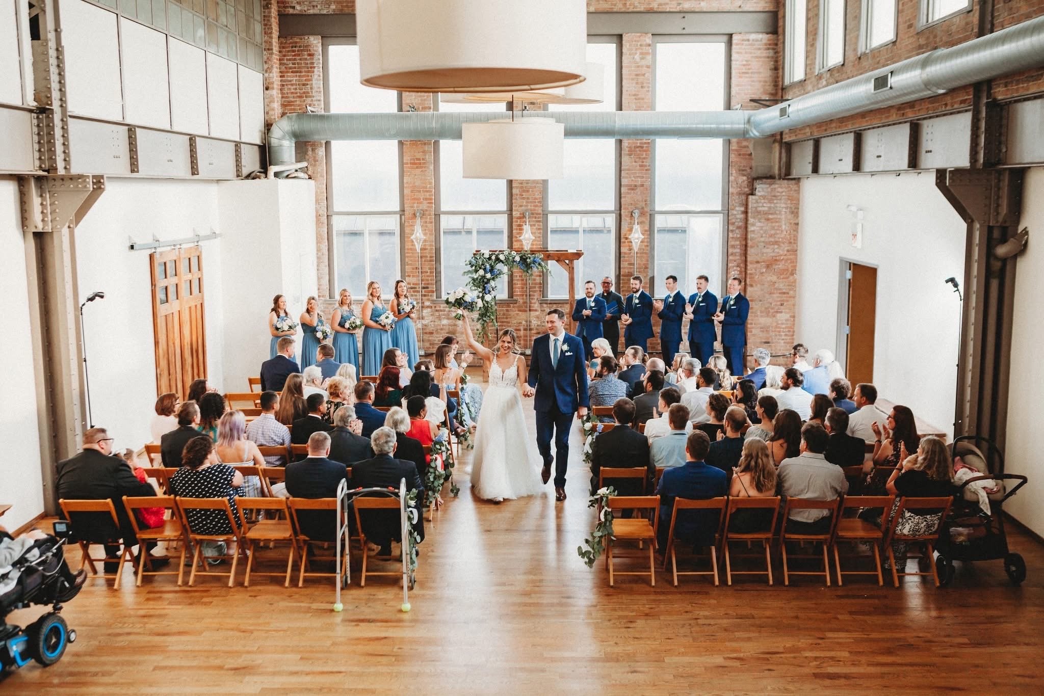 A wedding ceremony taking place in an industrial-style venue with brick walls and large windows, featuring a bride and groom walking down the aisle, surrounded by guests, with bridesmaids in blue dresses and groomsmen in matching suits on stage.