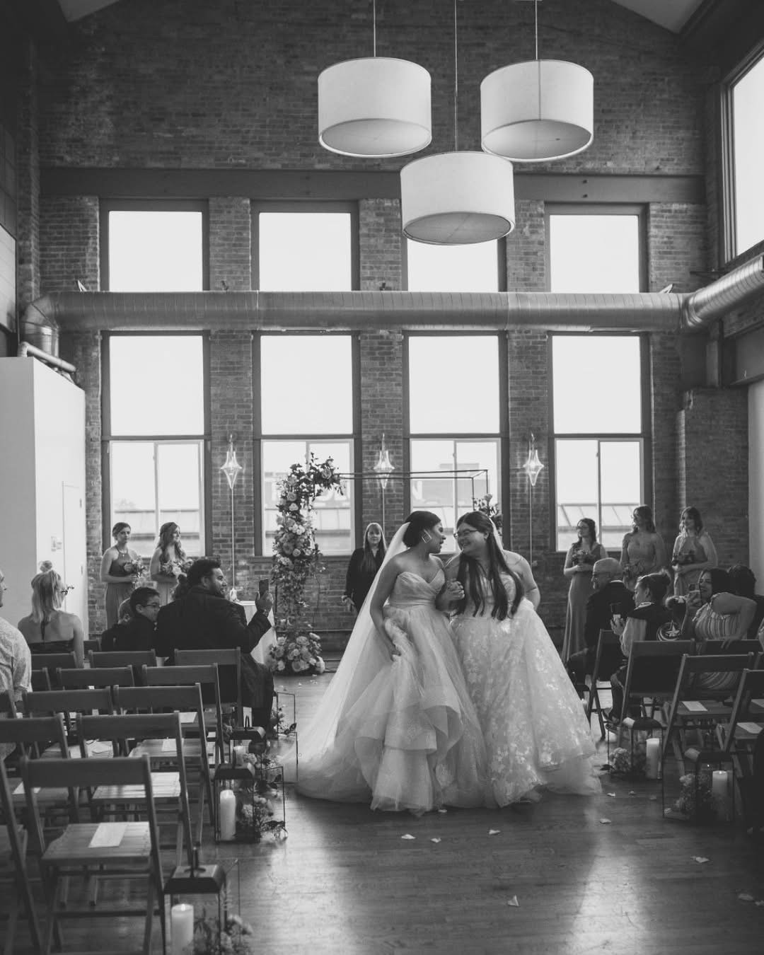 Black and white photo of a wedding ceremony in a modern venue with exposed brick walls and large windows. Two brides in wedding dresses are walking down the aisle, surrounded by seated guests and bridesmaids.