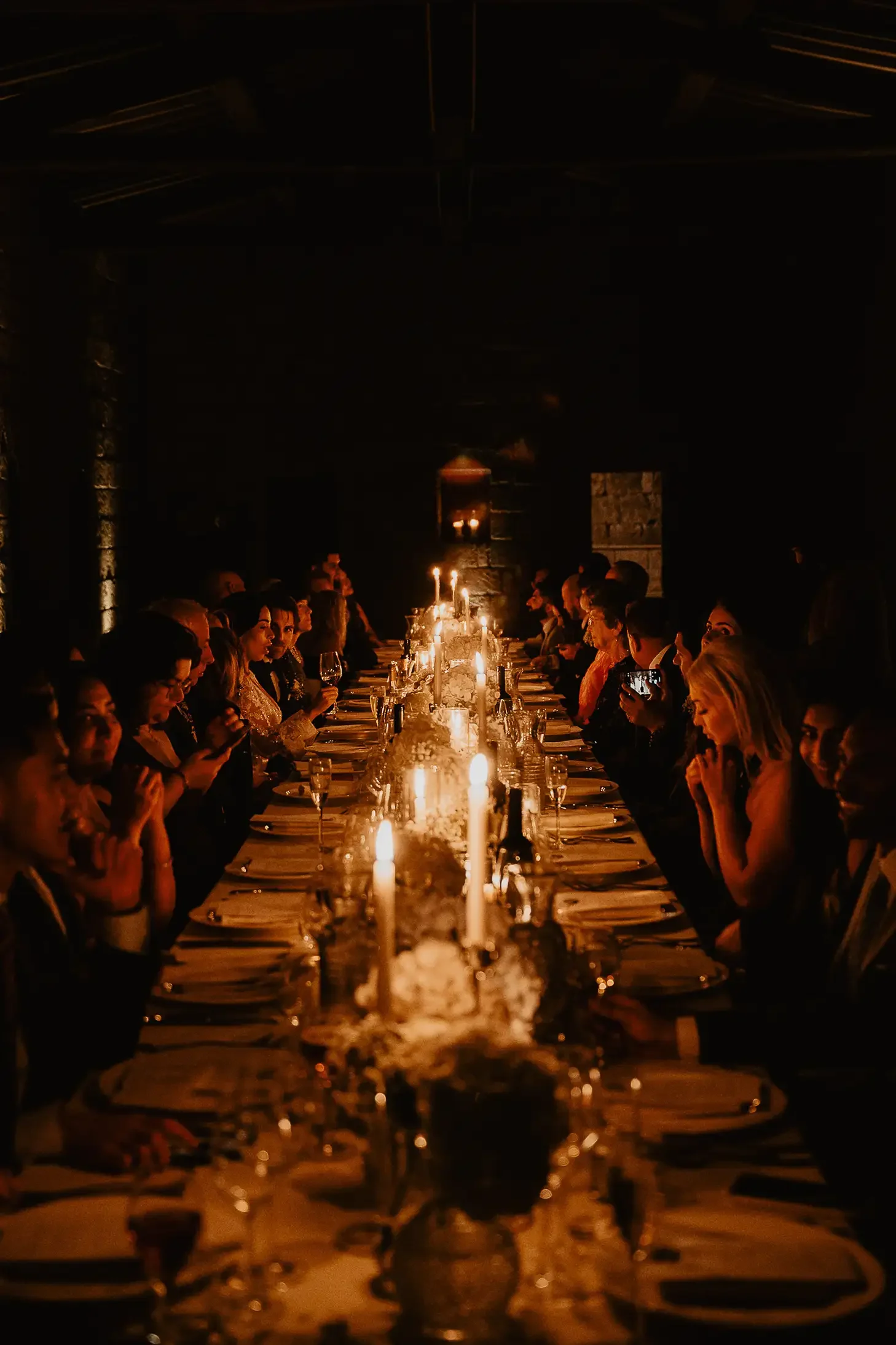A long dinner table in a dimly lit room with candles and floral centerpieces, surrounded by people dressed formally, during an evening event or celebration.