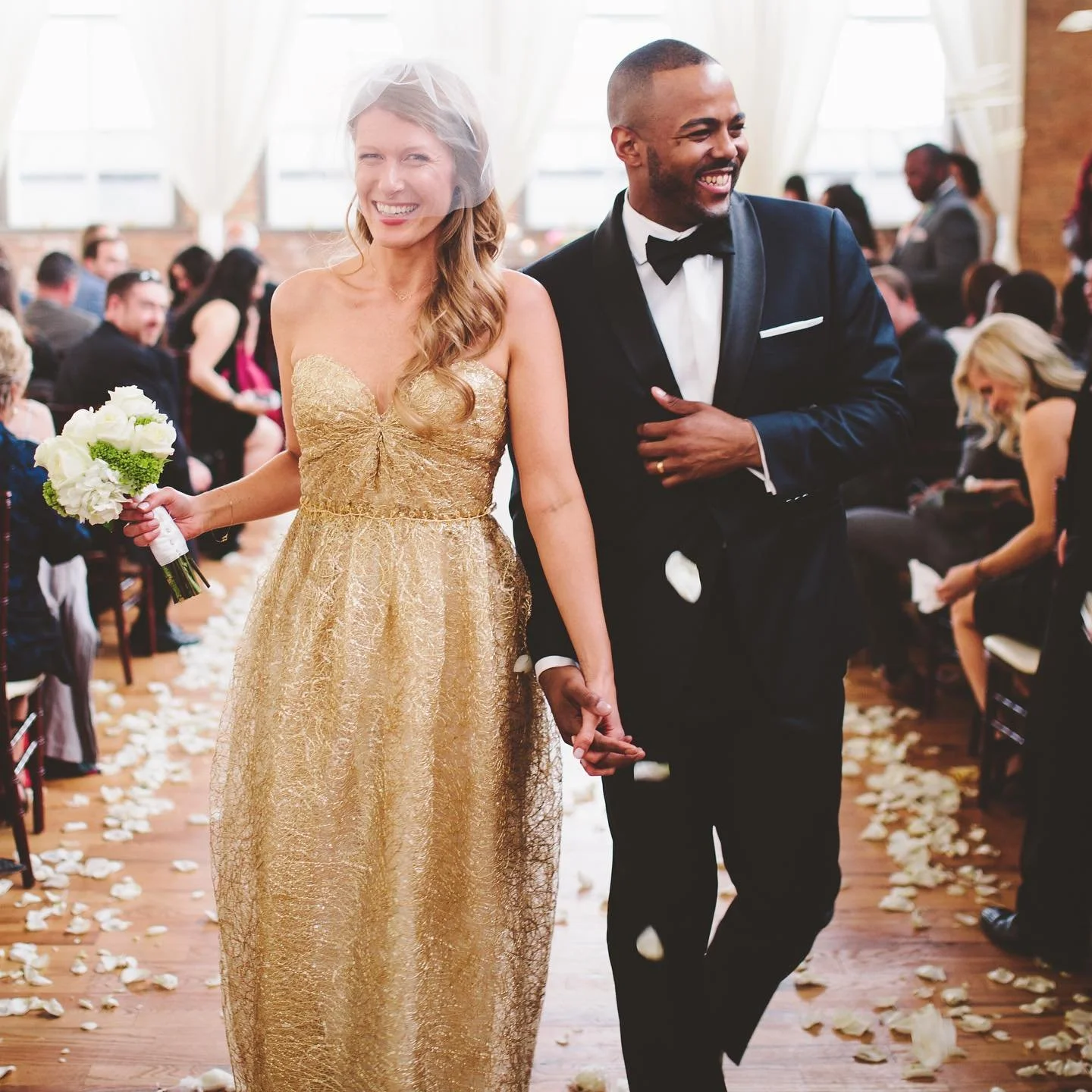 A bride and groom walking down the aisle at a wedding, smiling and holding hands, with guests seated on either side and flower petals on the floor.