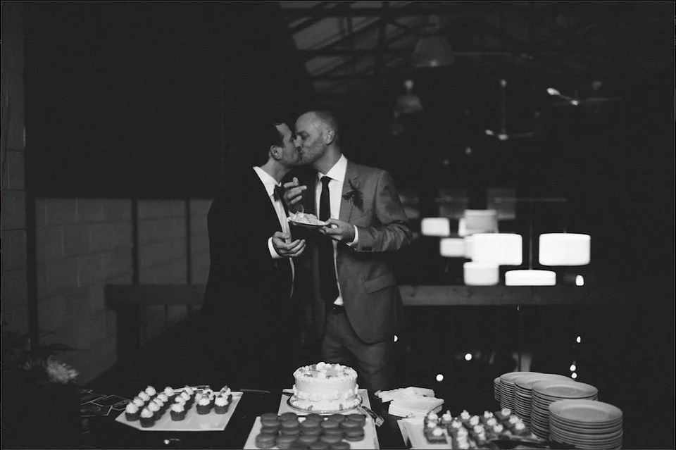 Two men in suits sharing a kiss at a wedding reception table with a cake and desserts.