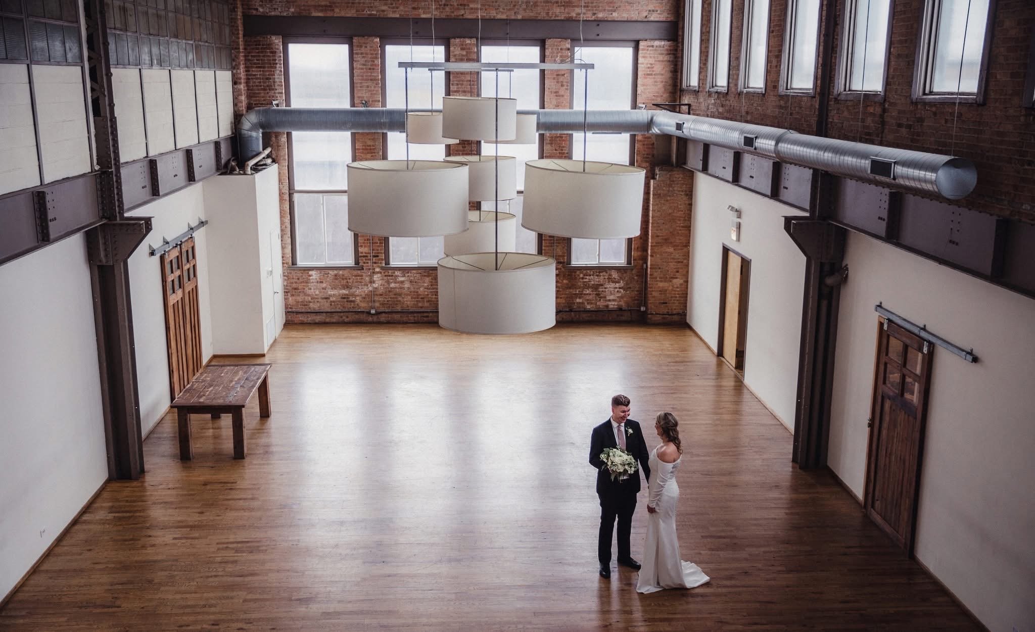 A bride and groom standing in an industrial-style venue with high ceilings, exposed brick walls, and large windows, sharing a handshake and looking at each other with a bouquet of flowers.