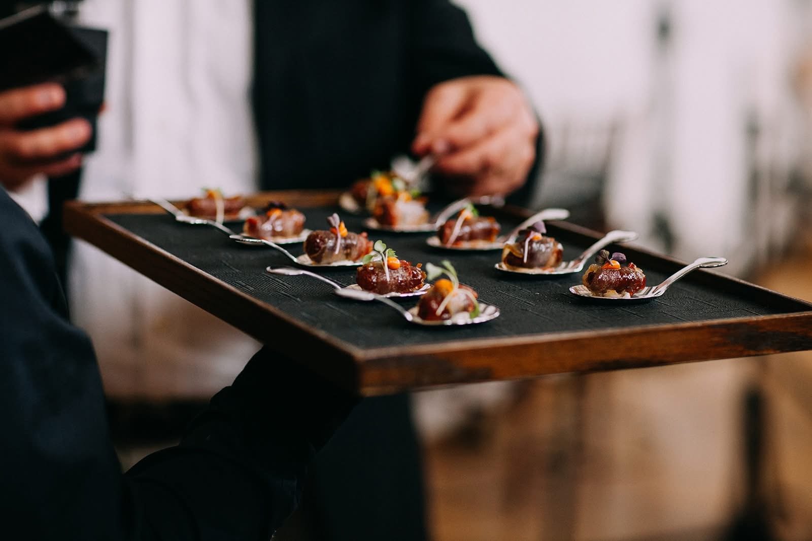 A server holding a tray with multiple small, gourmet appetizer bites on spoons, garnished with microgreens and colorful toppings.