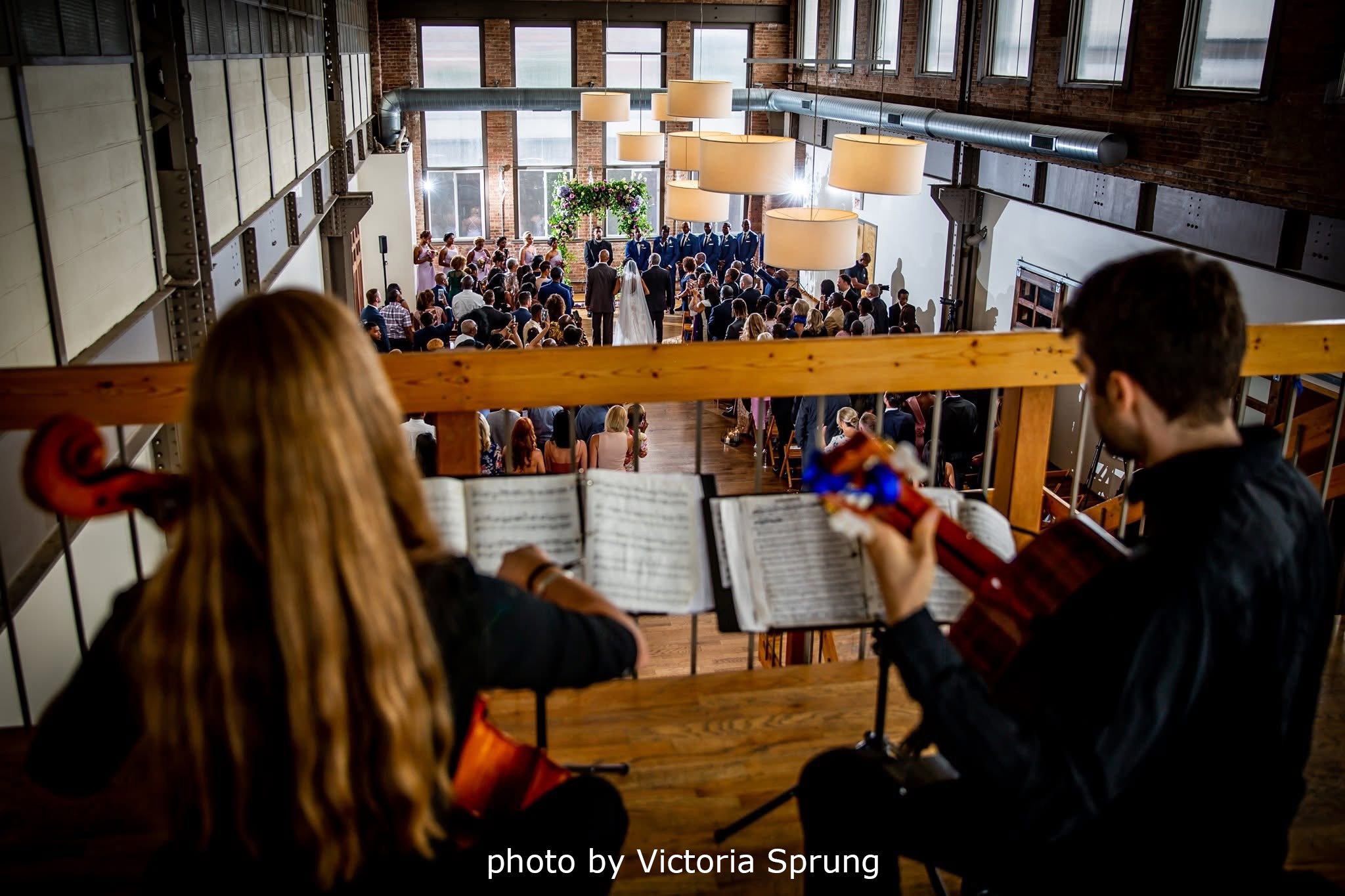 Musicians playing guitar and violin overlooking a wedding ceremony in a spacious industrial-style hall with large windows and hanging lamps.