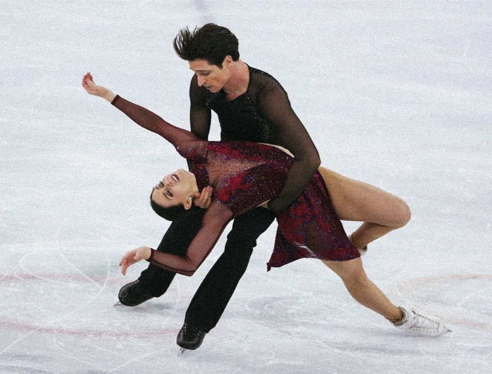A male and female figure skater performing a partner lift on an ice rink. The female is in a deep stretch with her head back and arms extended, while the male supports her from behind.