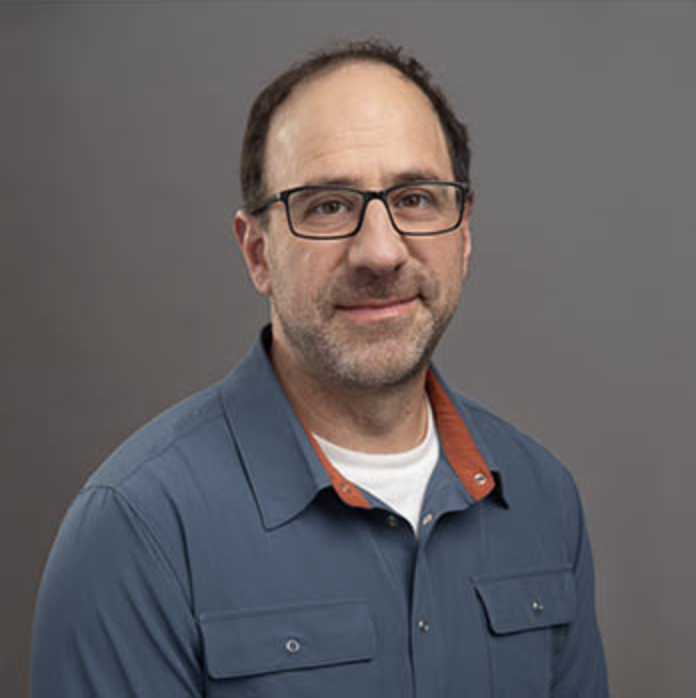 A middle-aged man with glasses, short dark hair, and a beard, wearing a blue collared shirt with an orange collar detail, standing against a gray background.