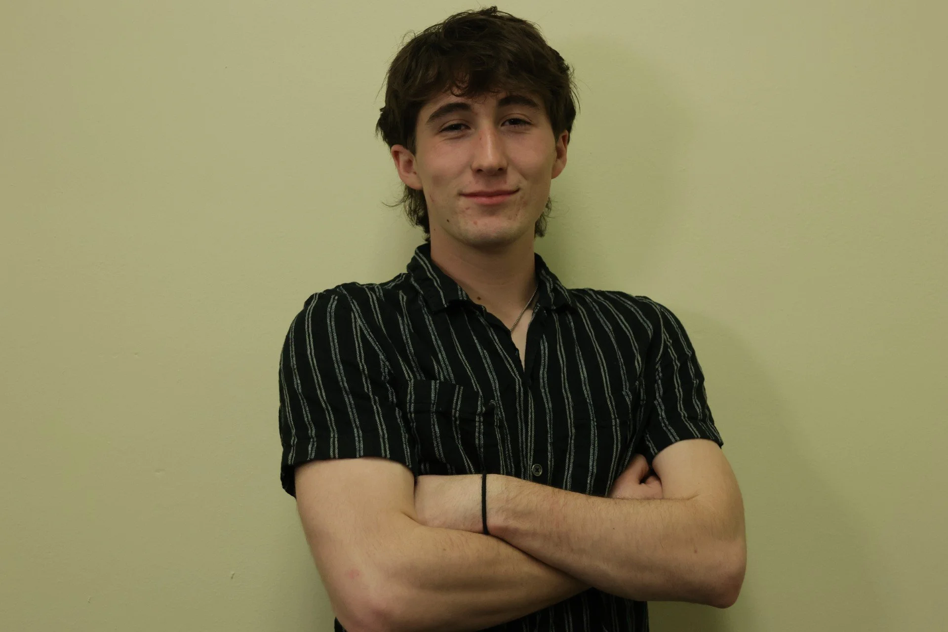 Young man with brown hair, wearing a black and white striped short-sleeve shirt, arms crossed, smiling slightly, standing against a plain yellow wall.