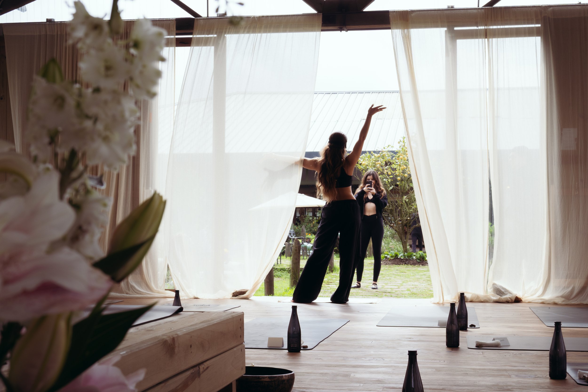 Woman with long hair dancing with one arm raised in a sunlit barn studio with sheer white curtains, another person filming her on a phone nearby, yoga mats and dark glass bottles on the wood floor.