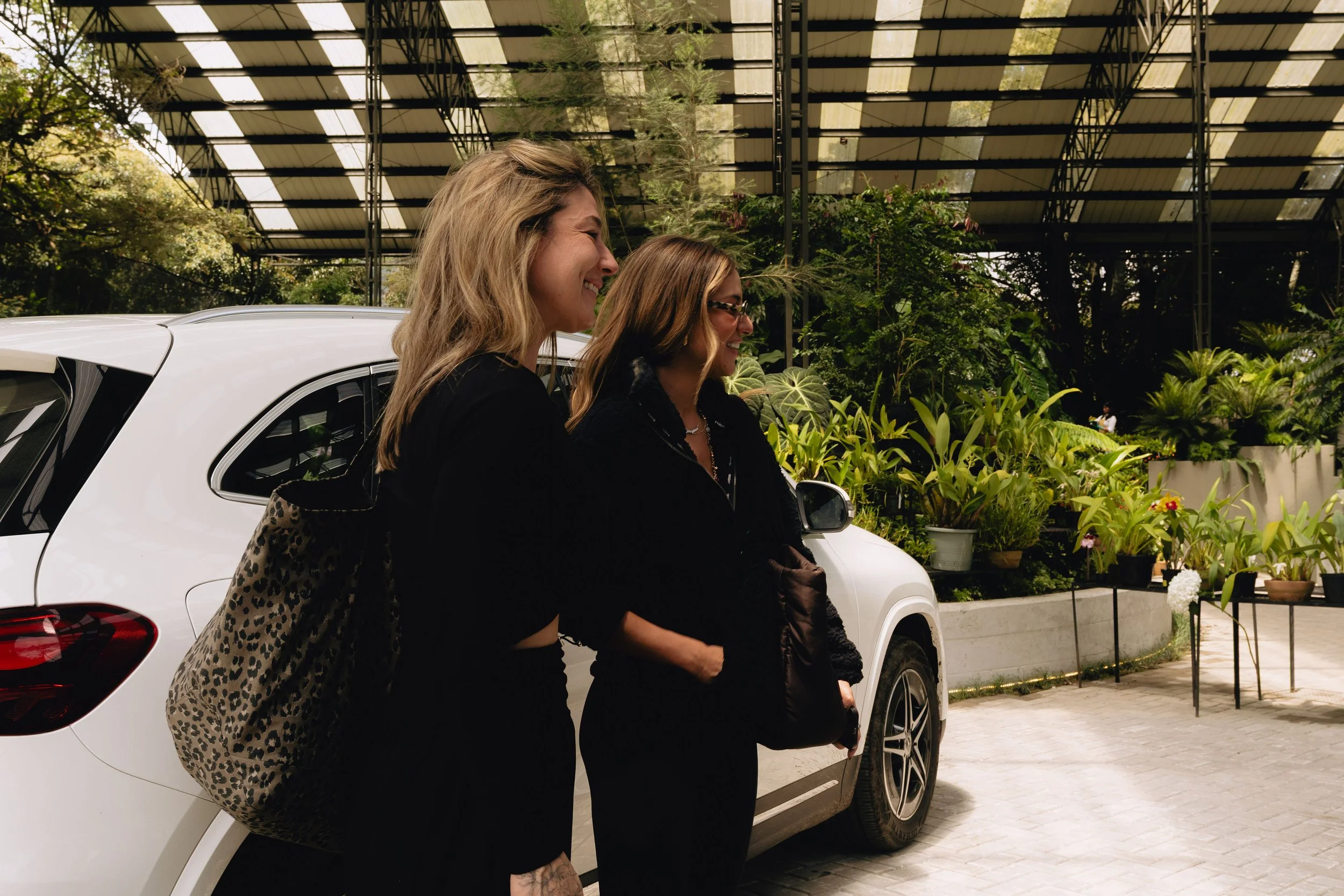 Two women laughing together beside a white car under a wooden pergola, one in a leopard-print jacket with wavy hair, the other in sunglasses with straight dark hair, both dressed in black, surrounded by tropical potted plants on a paved driveway.