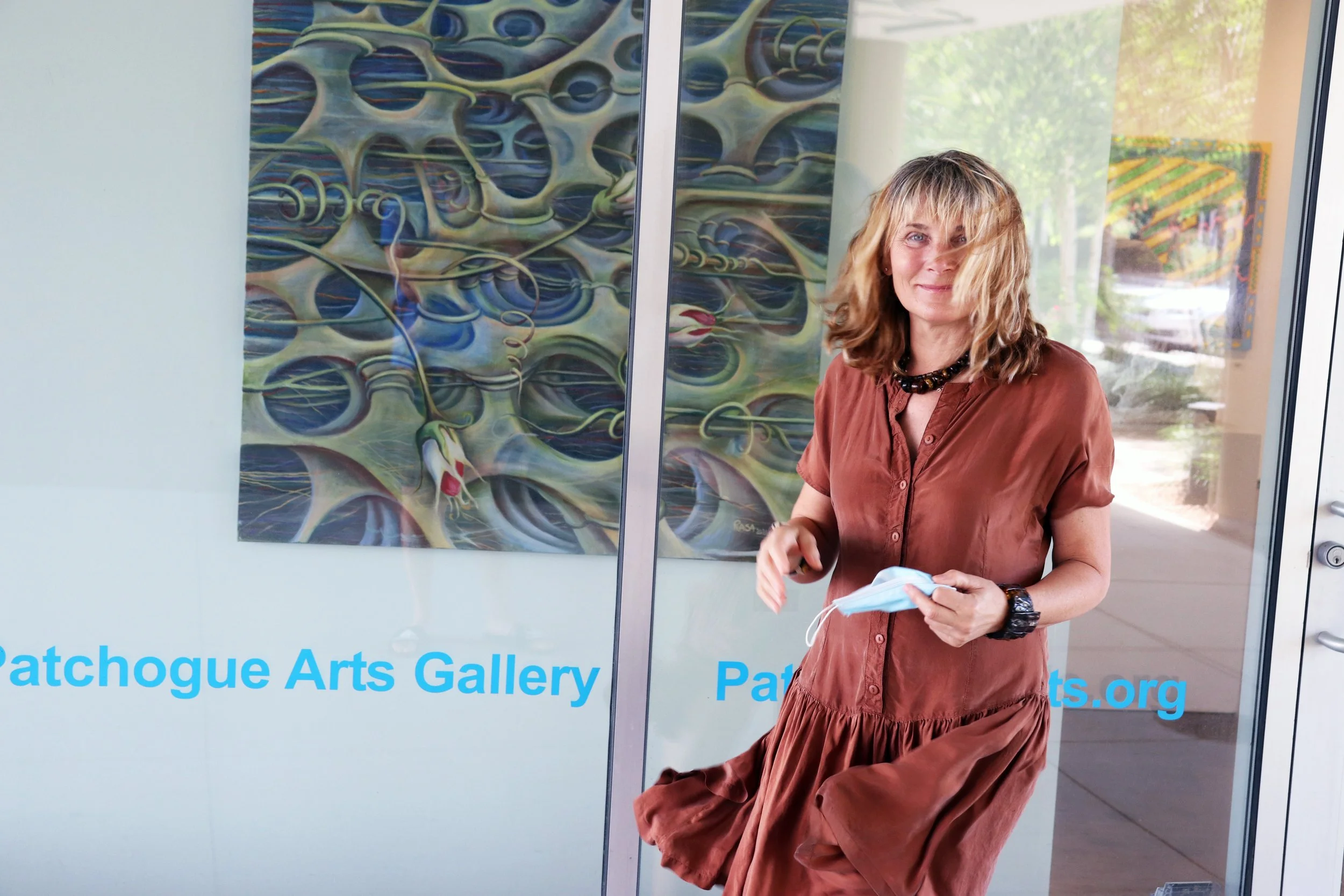A woman with shoulder-length blonde hair, wearing a rust-colored dress, stands outside a glass door with a painting of abstract interconnected shapes behind her. She holds a face mask in her right hand and smiles at the camera. The glass door has the text 'Patchogue Arts Gallery' and the website 'PatchogueArts.org' in blue.