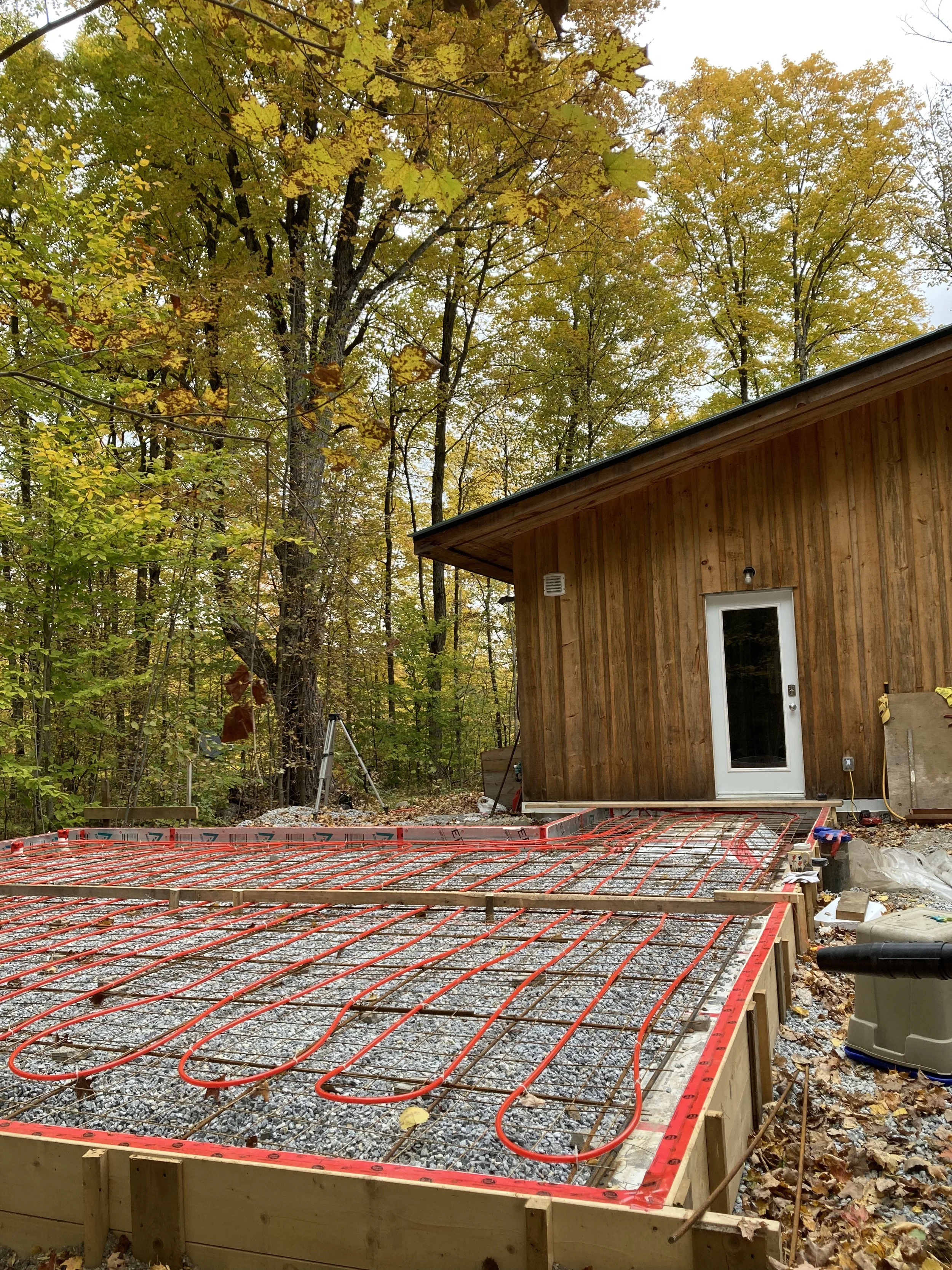 Construction site showing a wooden house with a glass door and a partially built concrete slab with embedded red tubing for radiant floor heating, surrounded by trees with autumn foliage.