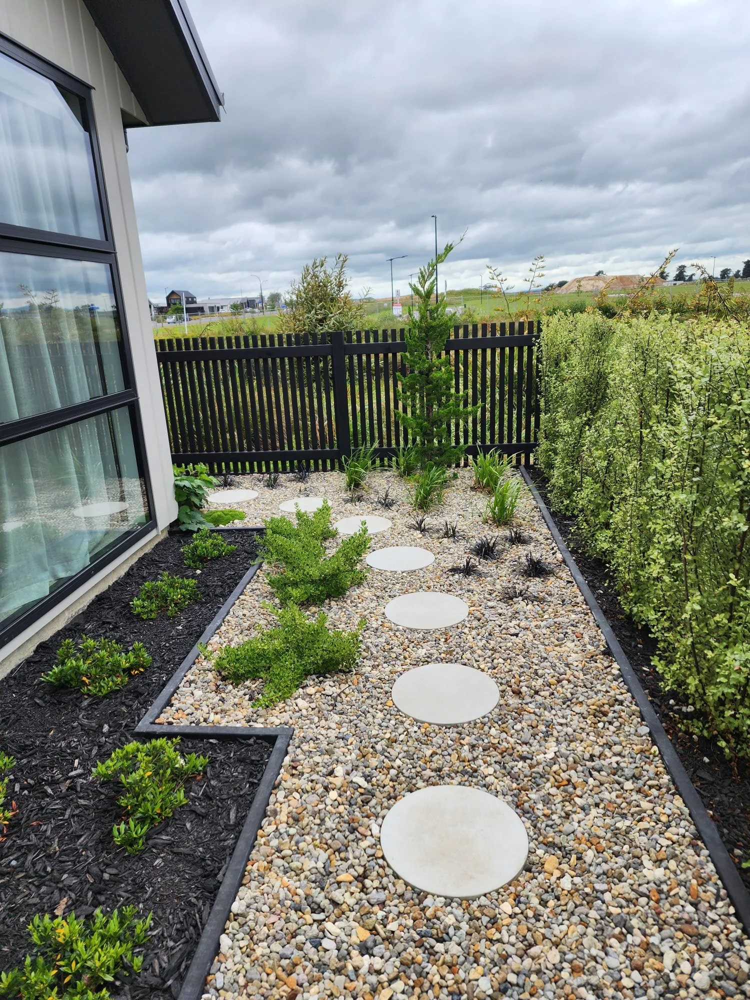 A landscaped backyard with a stone path featuring round stepping stones, bordered by black edging, with small shrubs and plants on both sides, and a black wooden fence in the background. The sky is overcast with gray clouds.