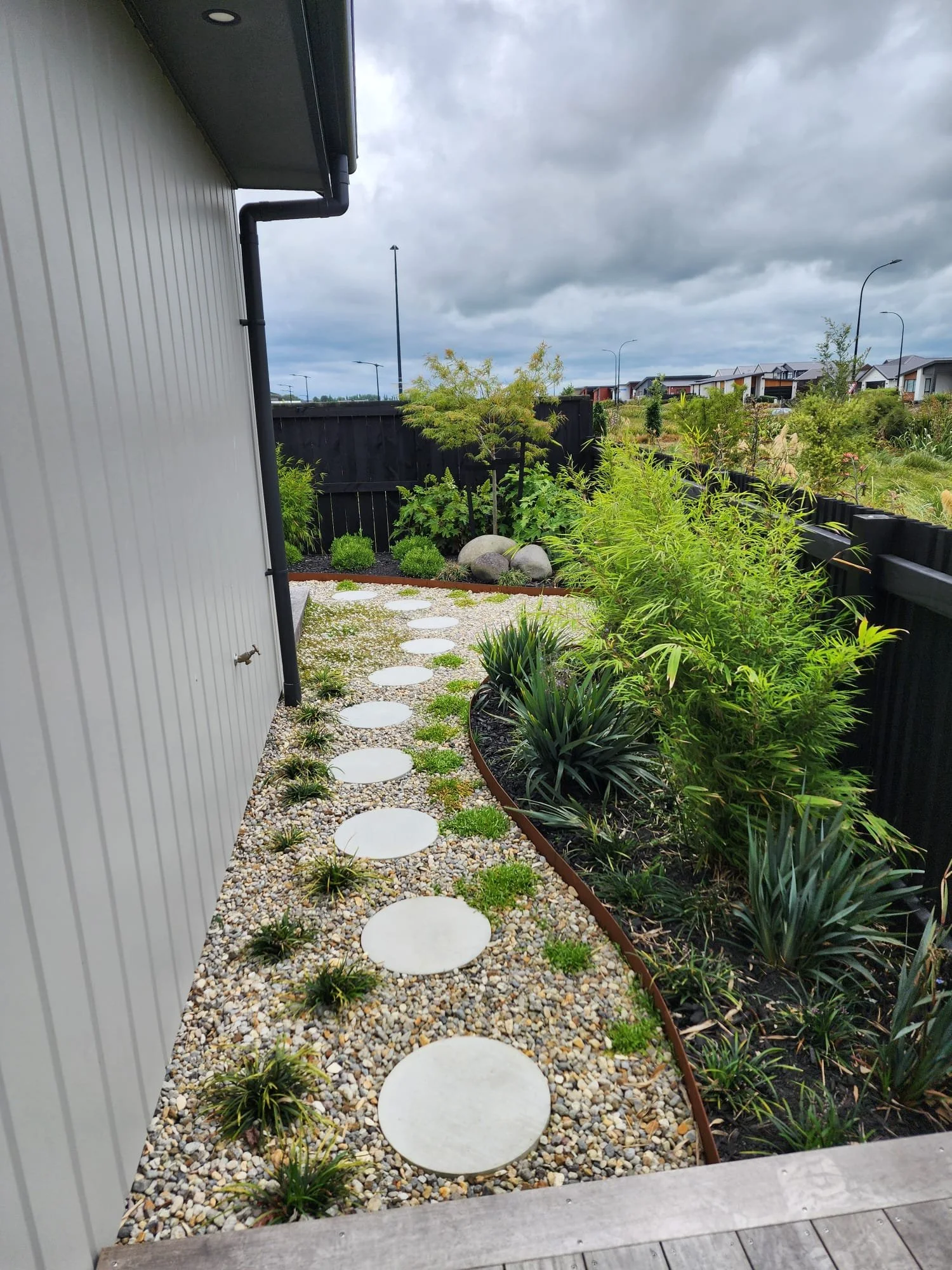 Side garden with a curved pathway made of round white stepping stones and small plants, bordered by a metal trim, adjacent to a house with gray siding and black gutter, with lush green shrubs and trees, fence, and street lights in the background unde