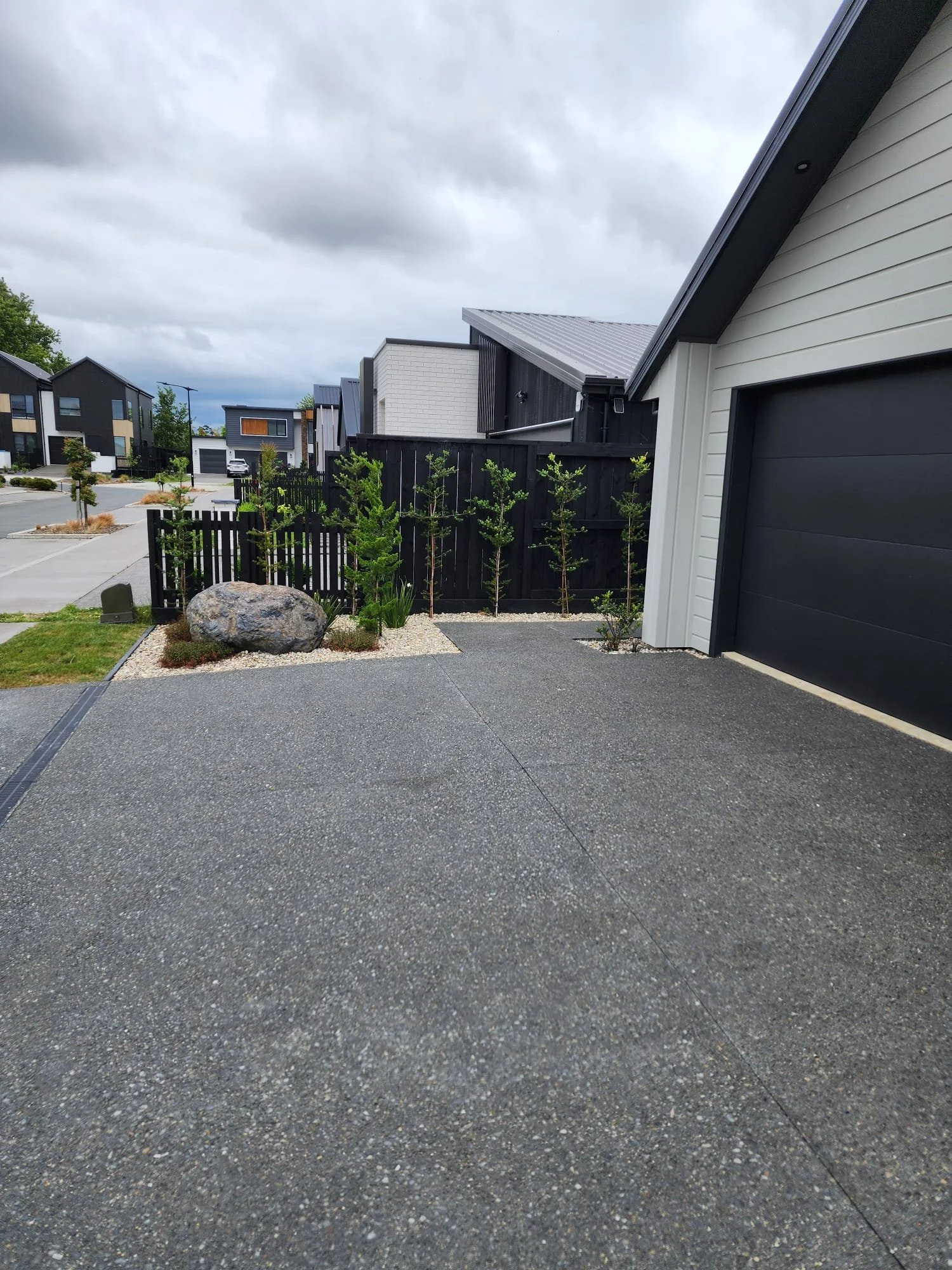 Residential driveway with a large rock and small trees bordered by white gravel; modern houses with black and white exteriors in the background under cloudy sky.