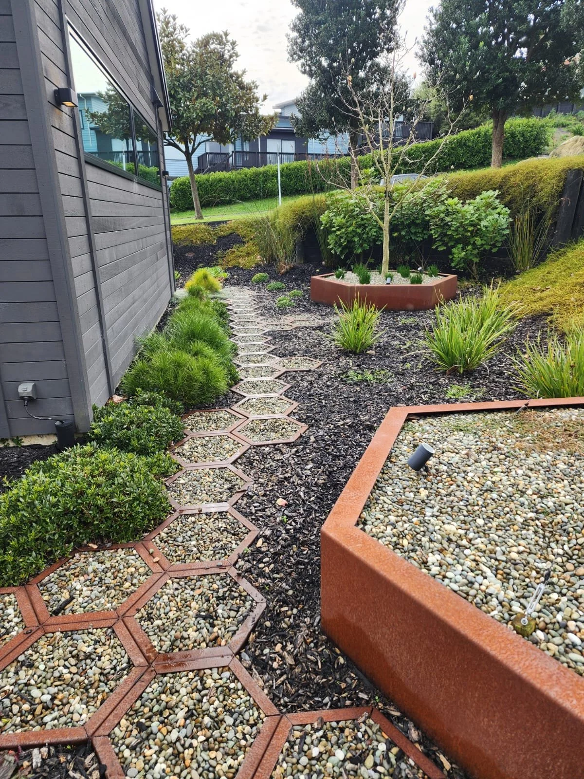 View of a modern backyard garden with a stepping stone path, shrubs, a small tree in a border, and a raised gravel bed near a gray house with dark siding.
