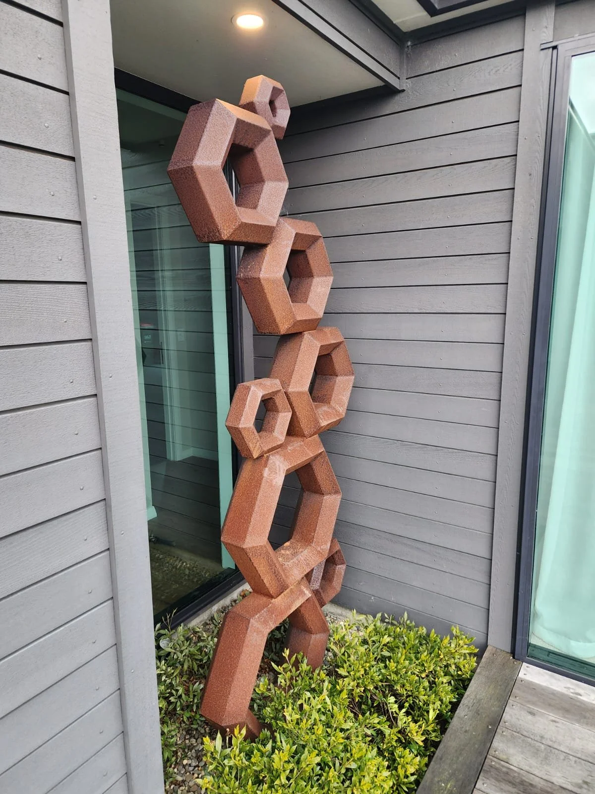 An outdoor decorative wooden sculpture with interconnected geometric shapes, situated against a house with gray siding and a sliding glass door, surrounded by green shrubs.