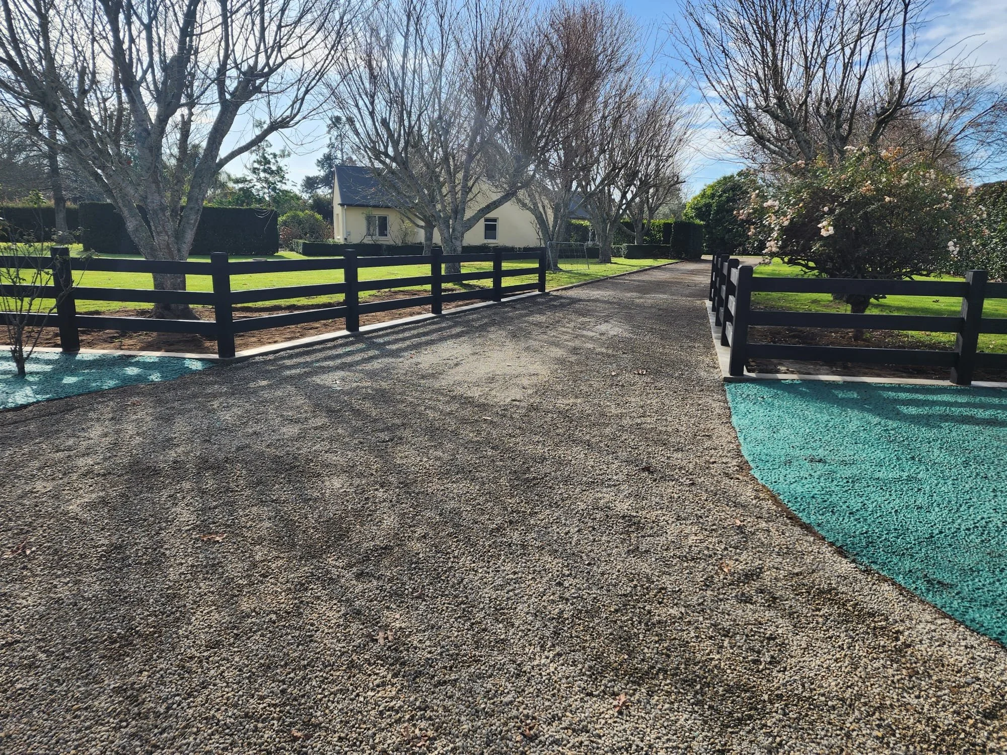 A gravel driveway leading to a house with a cream exterior and dark roof, flanked by trees and bushes, some with blossoms, and bordered by black fencing, under a partly cloudy sky, with sunlight casting shadows.