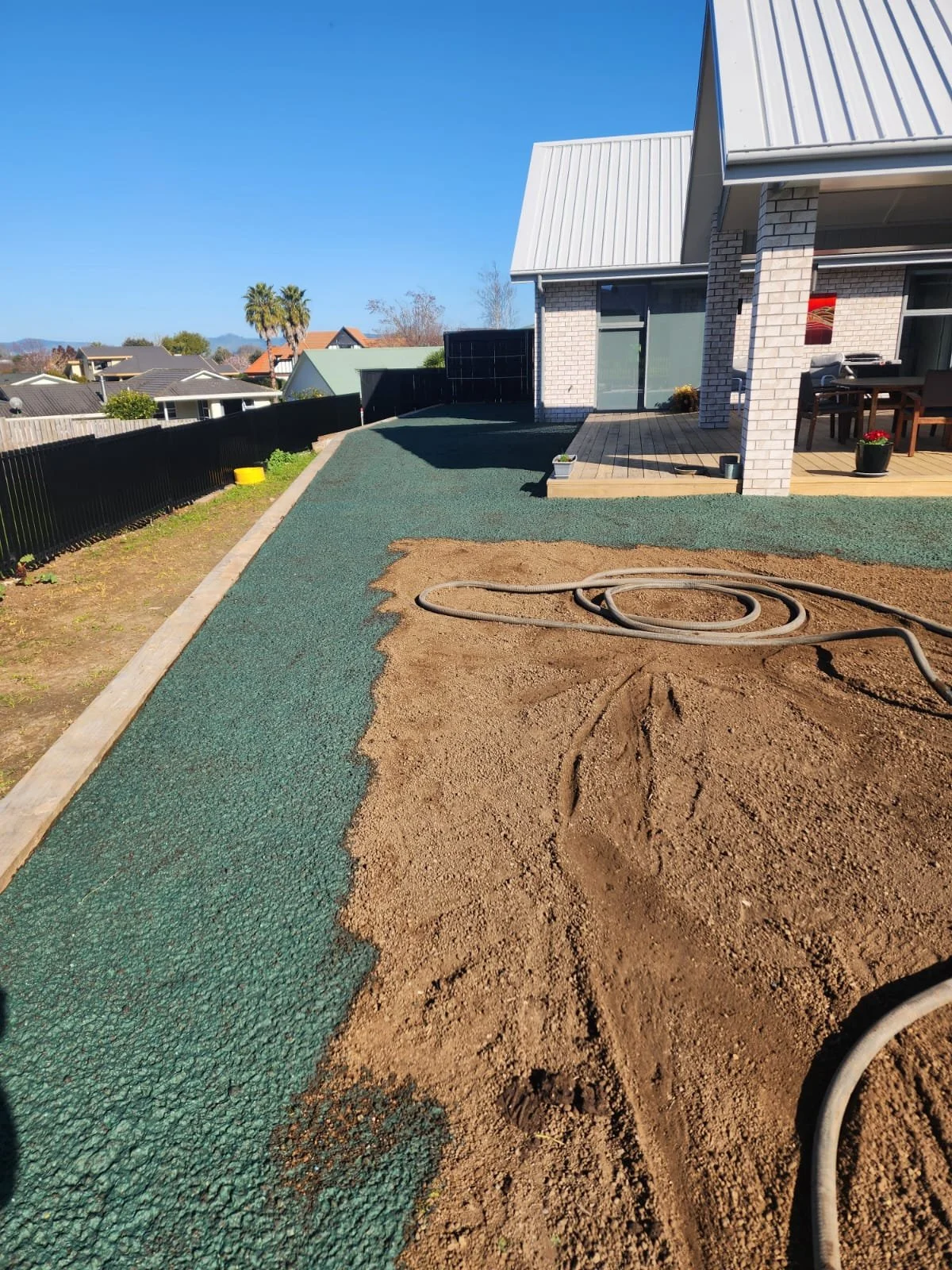 Backyard under construction with a partially completed pathway, green rubberized surface, and exposed dirt area with hoses, in front of a house with a patio and outdoor furniture.