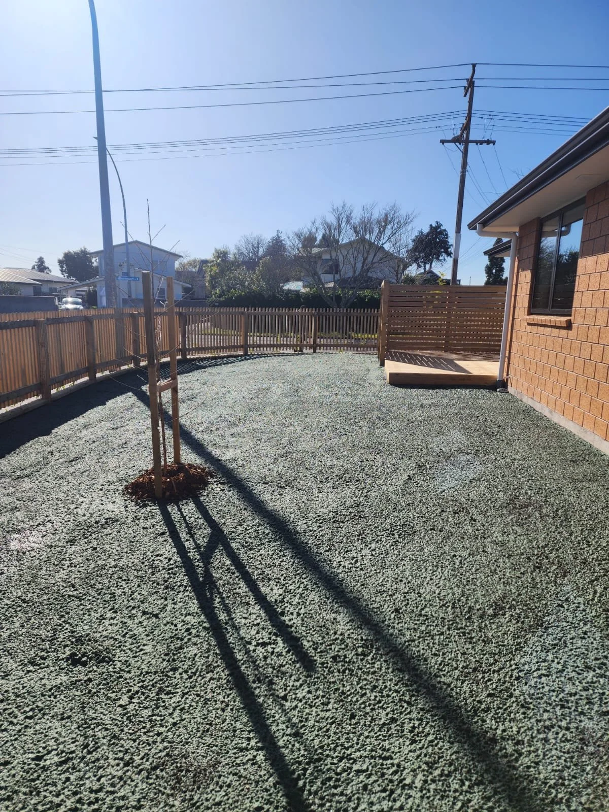 A backyard with a small newly planted tree, surrounded by a wooden fence. The ground is covered with gravel, and there is a wooden deck attached to a brick house on the right. Utility poles and power lines are visible in the background under a clear 