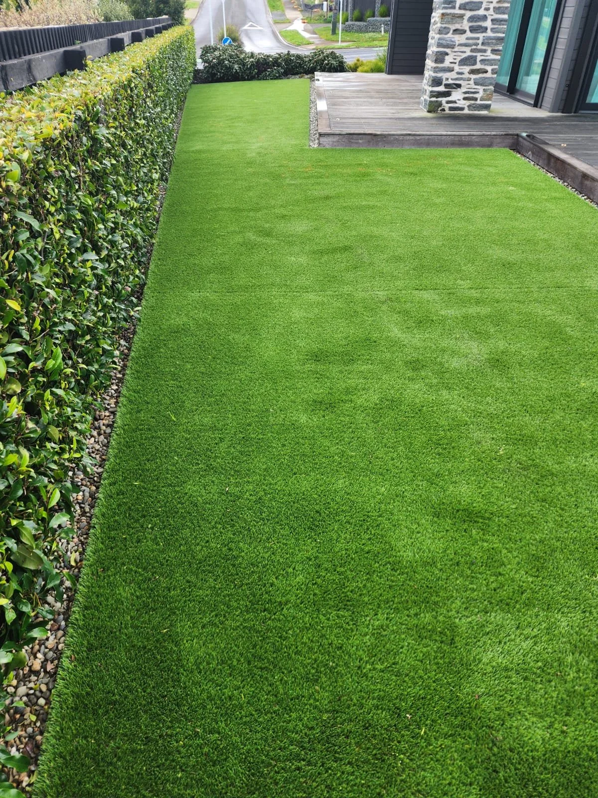 A well-maintained yard with a lush green artificial grass lawn, bordered on one side by a neatly trimmed hedge and on the other by a wooden deck and a stone house exterior.