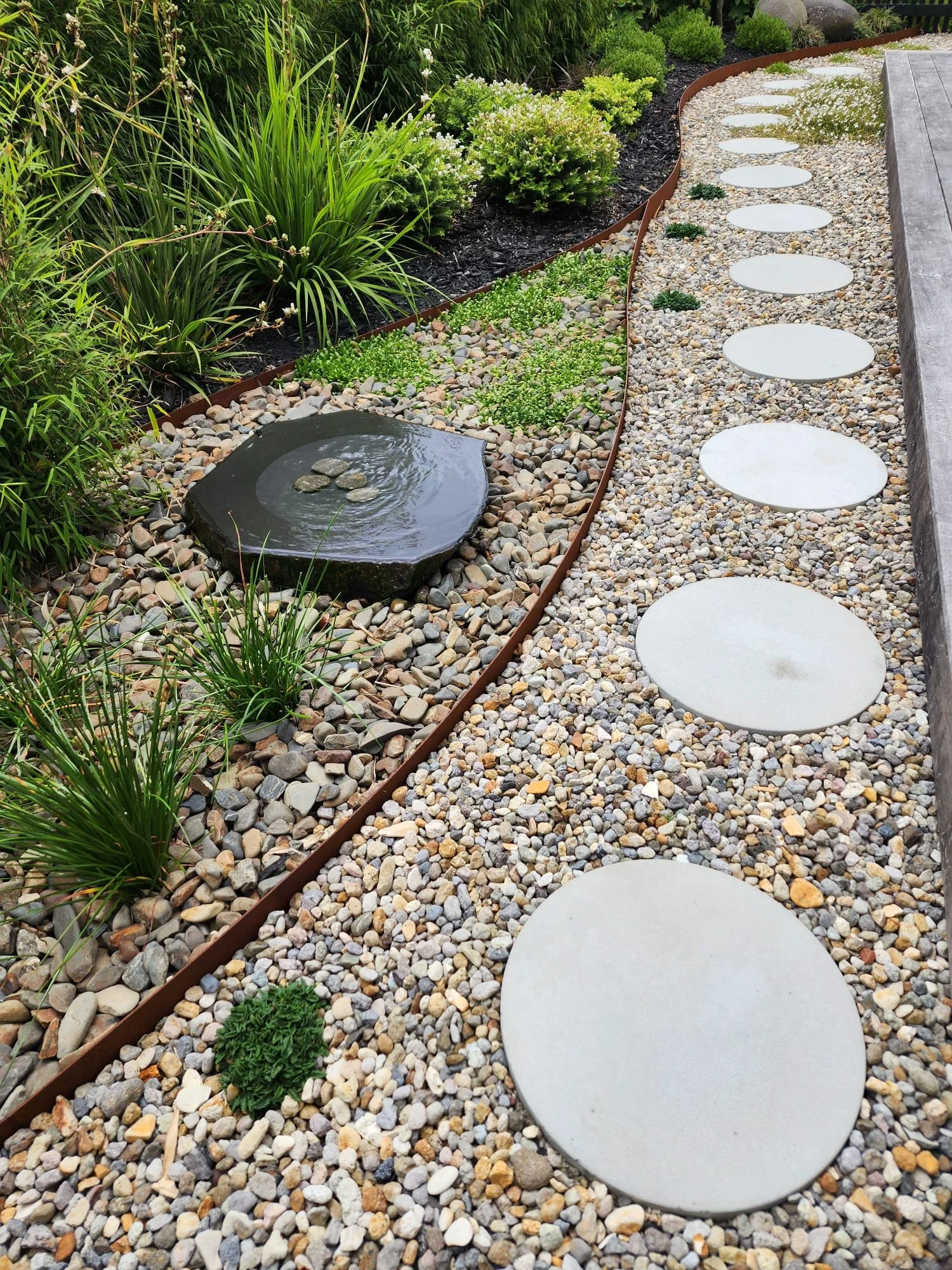 A landscaped garden pathway with round concrete stepping stones on a bed of small multicolored pebbles, bordered by a mixture of green plants and bushes, and a small water feature with rocks.