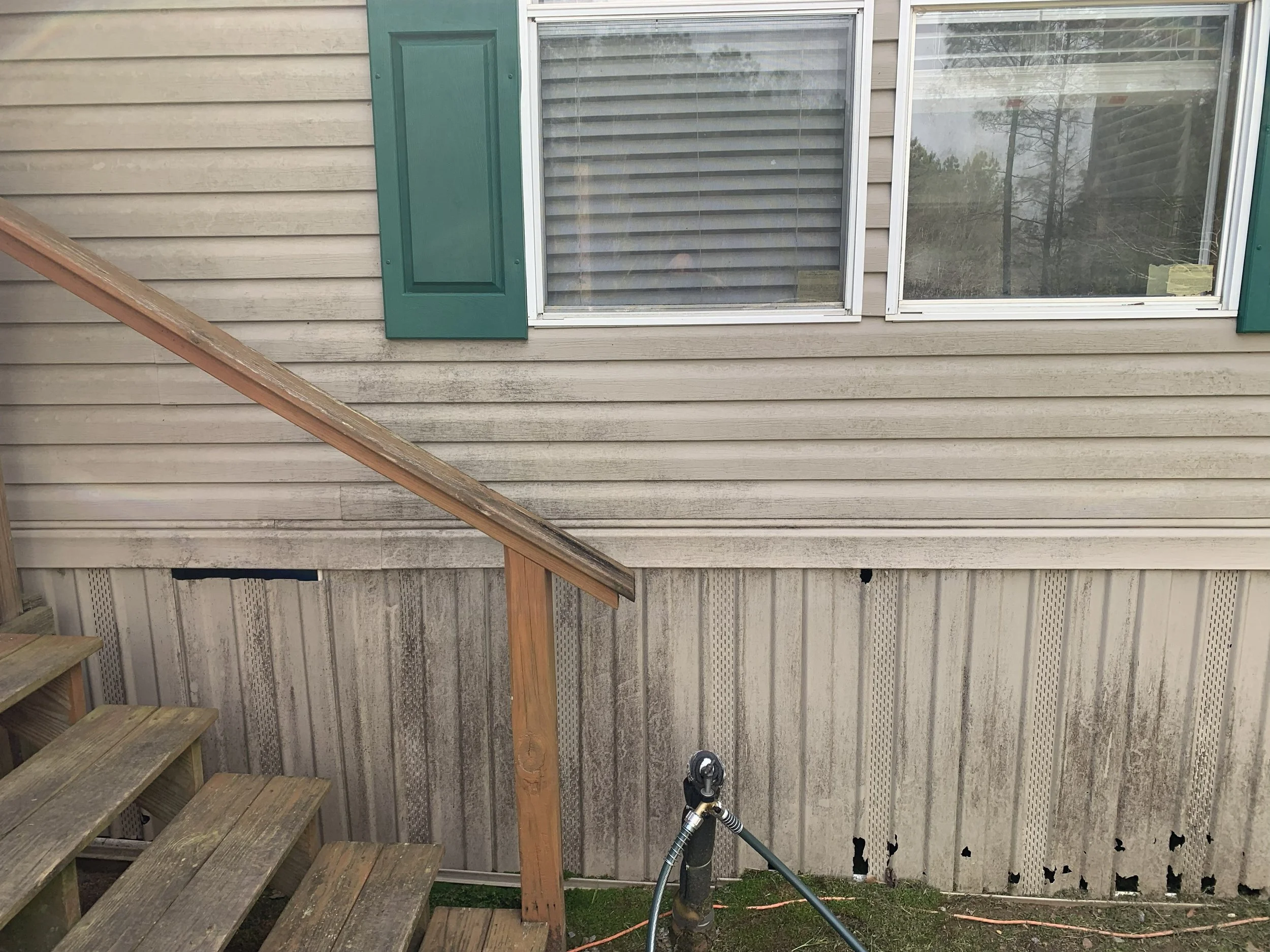 Backyard wooden deck with stairs on the left, adjacent beige siding house with two windows with green shutters, a garden hose on the ground, and a corrugated metal fence at the bottom of the image.