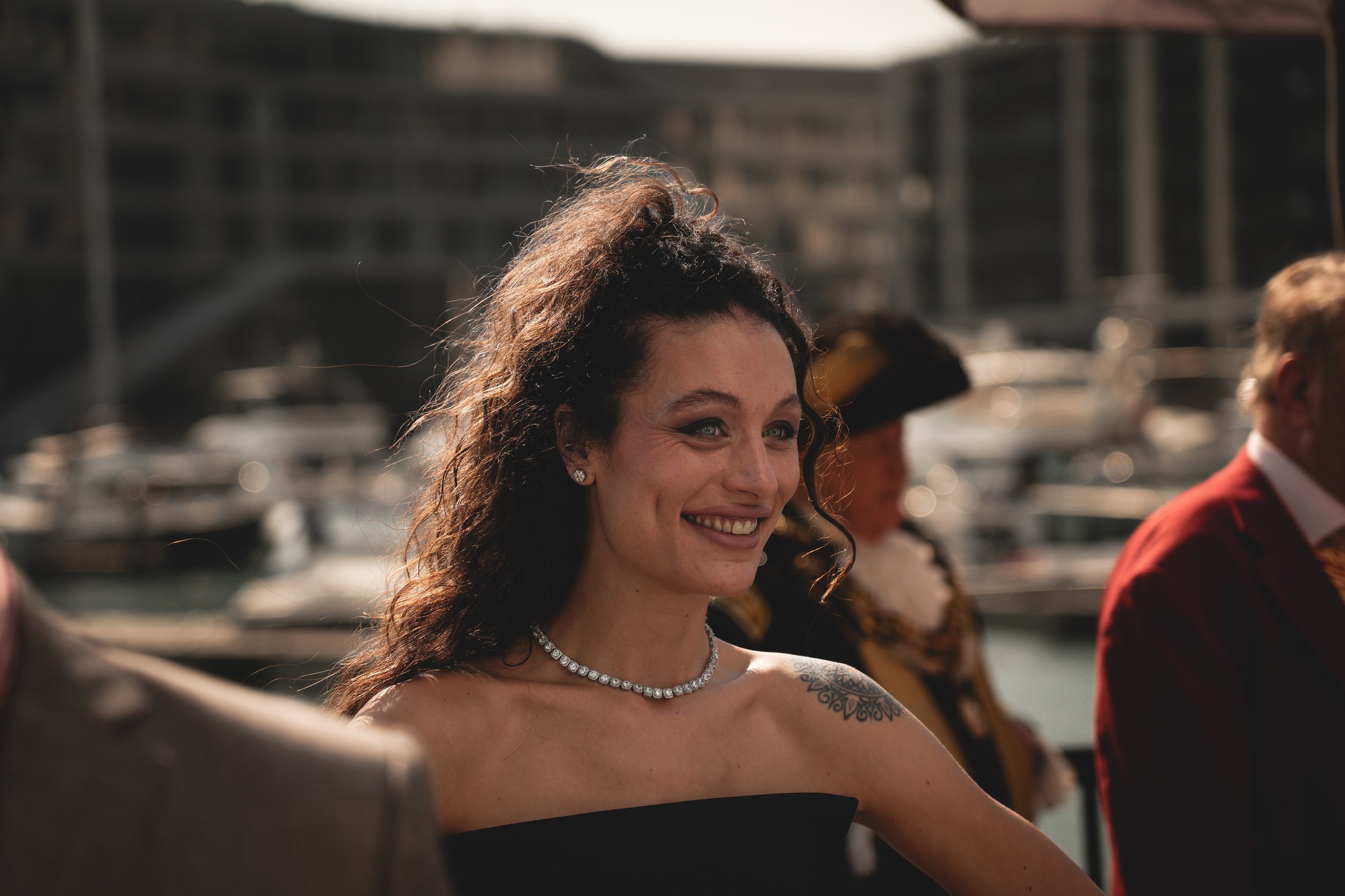 A woman with curly brown hair styled half-up, smiling at an outdoor event near a marina with boats, wearing a black dress, a pearl necklace, and diamond earrings.